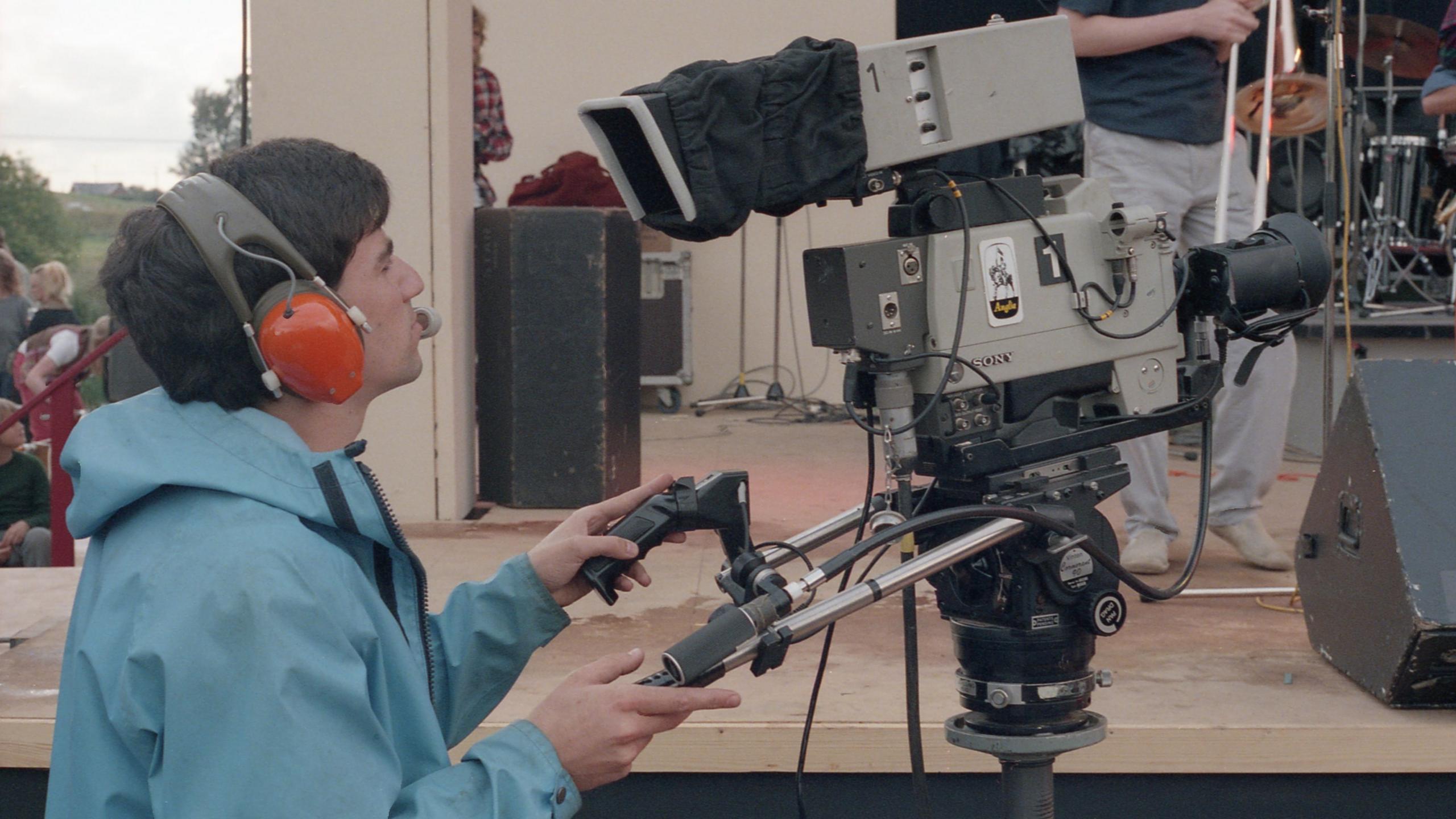 A colour mid-shot side view of a camera operator and a television camera, outside in daylight just in front of a large stage. The cameraman appears to be approximately in his 30s, has dark hair, and is seen left of frame wearing red headphones and a pale blue rain jacket. He is holding the operating handles of his large television camera and looking into its viewfinder. The camera is fixed onto a mounting and has an Anglia Television logo on its side.