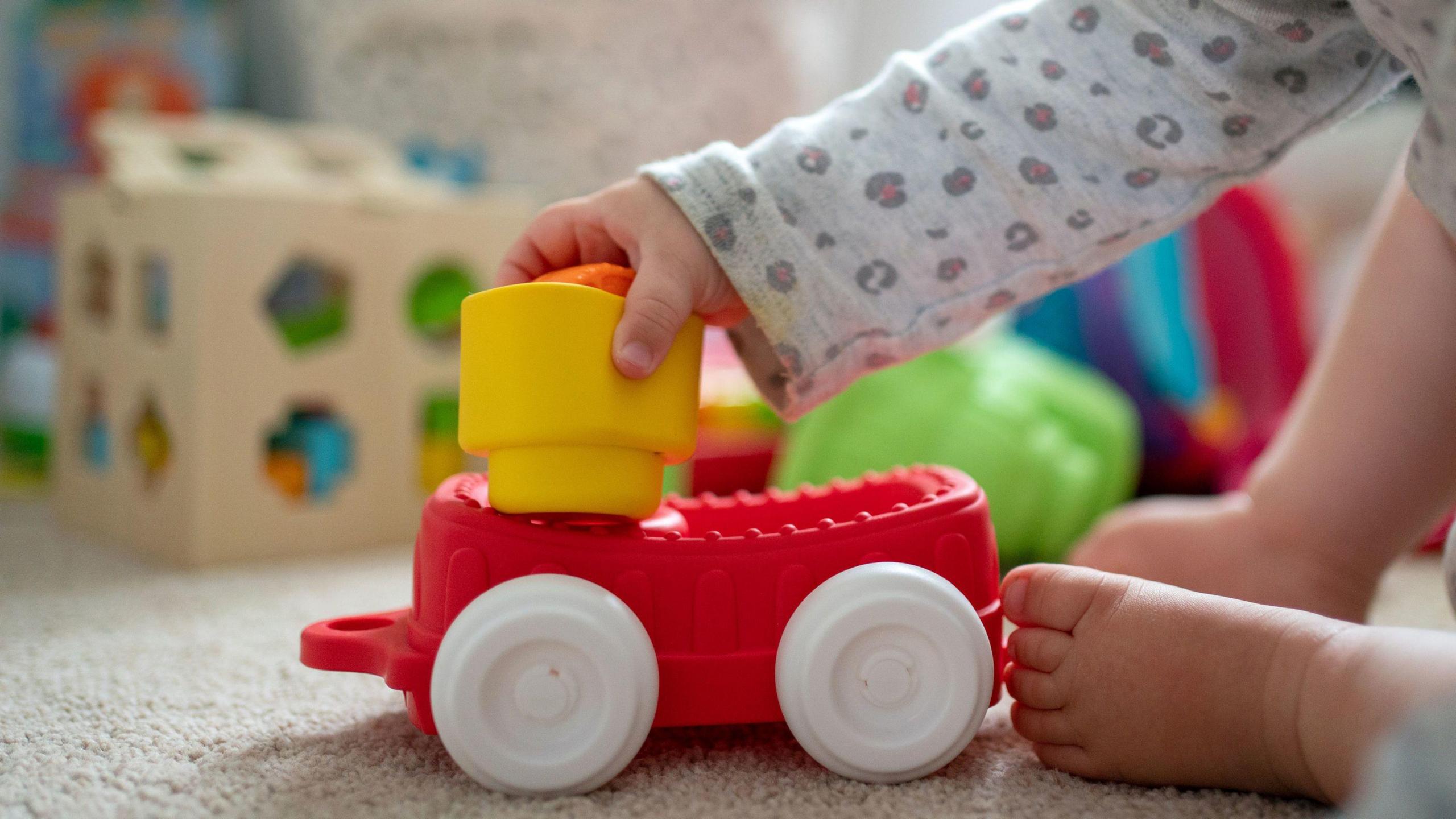 A baby holding a toy car. Only arm and feet are in shot, with blurred toys in the background.