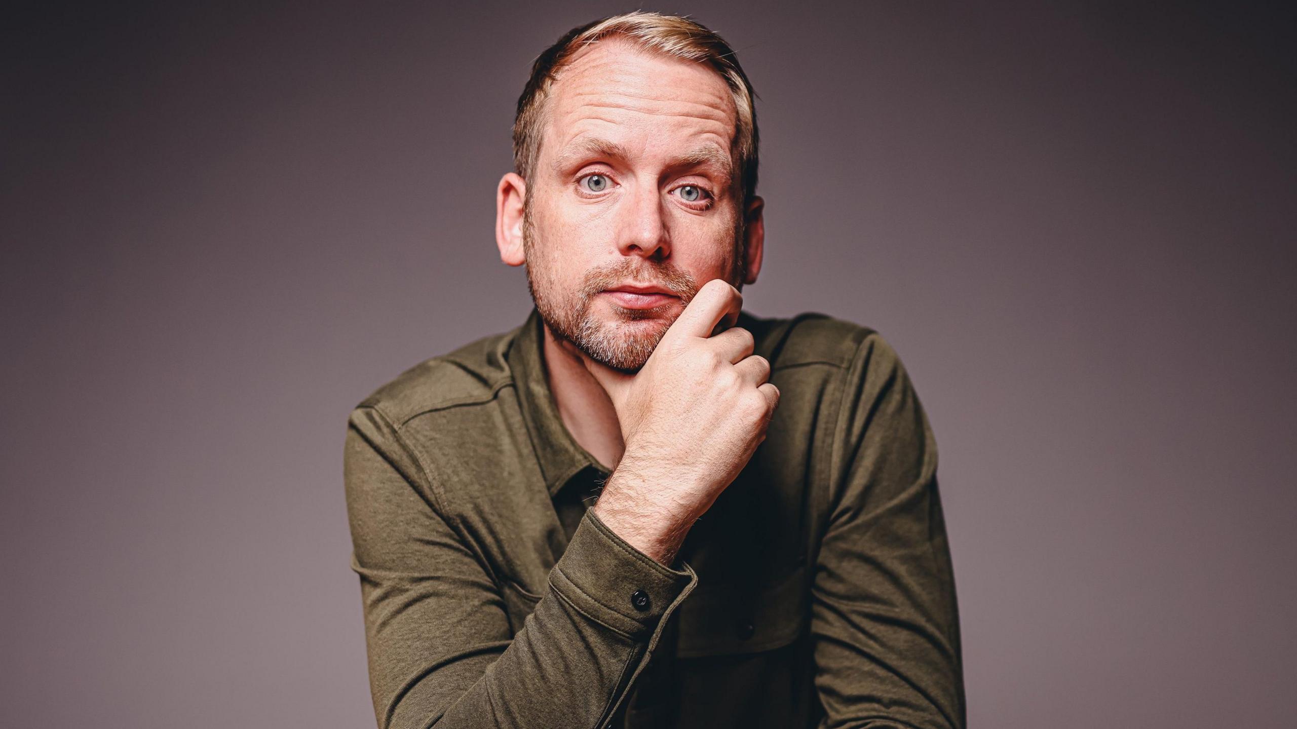 Andi looks at the camera with an inquisitive expression, resting his chin on one hand. He has short blond hair, a stubbly beard and wears a long sleeve dark green top. 