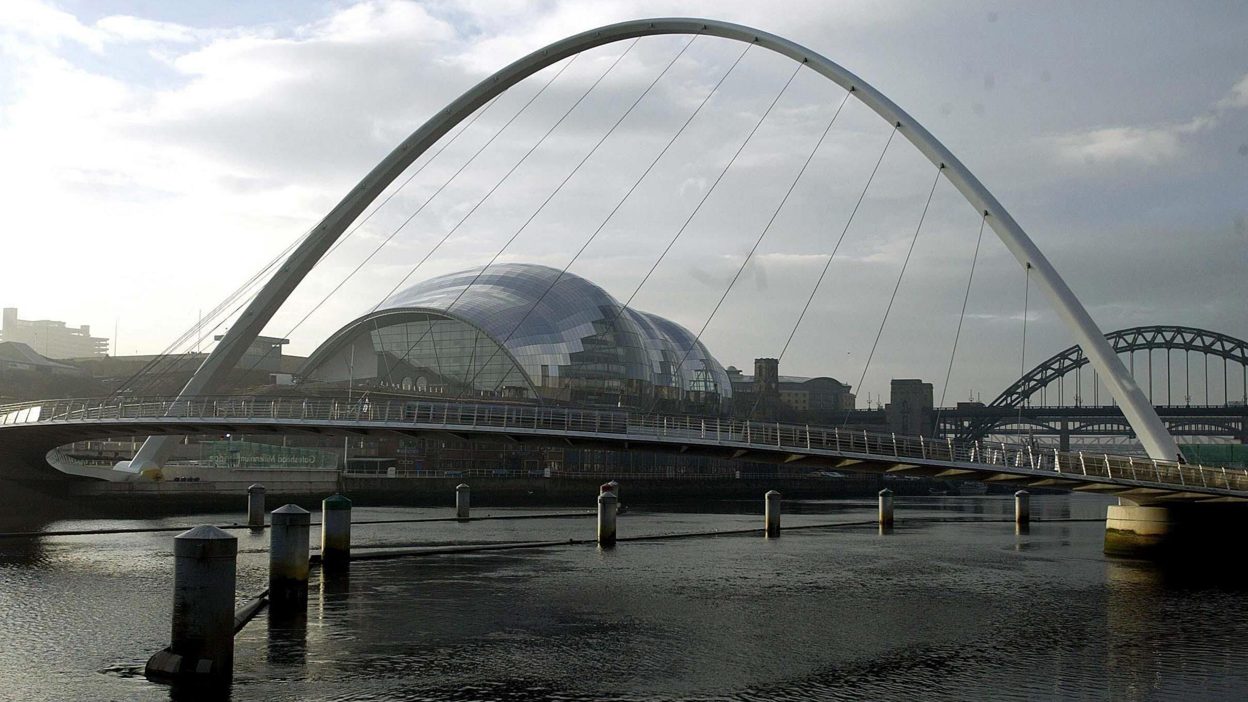 The Glasshouse which is a curved, glass building on the side of the River Tyne. The white Millennium Bridge can be seen in front of it and the green Tyne Bridge behind it.