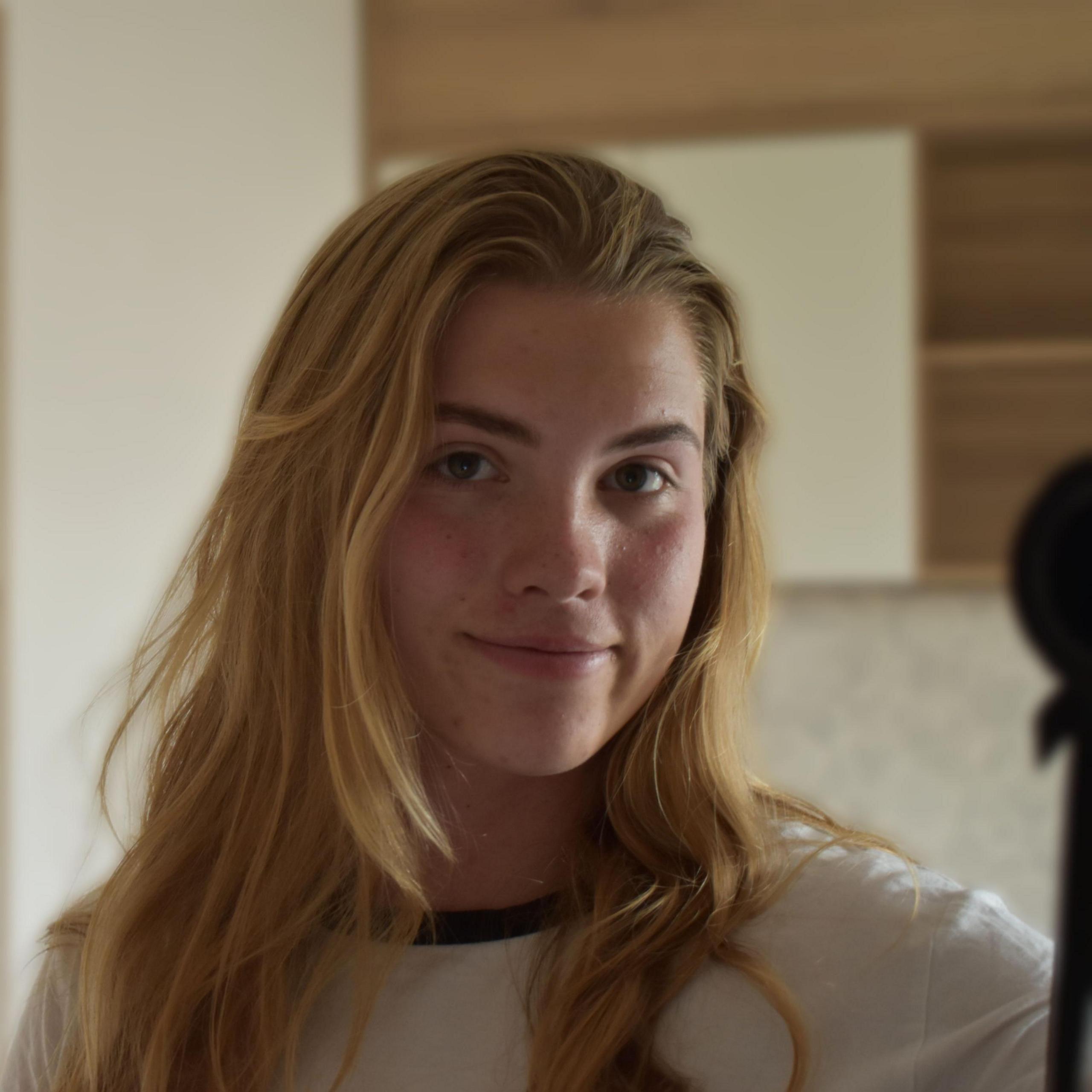 A self-portrait of Erin Gailey. She has long, light hair past her shoulders and is wearing a white top with a dark trim around the neck and slightly smiles at the camera