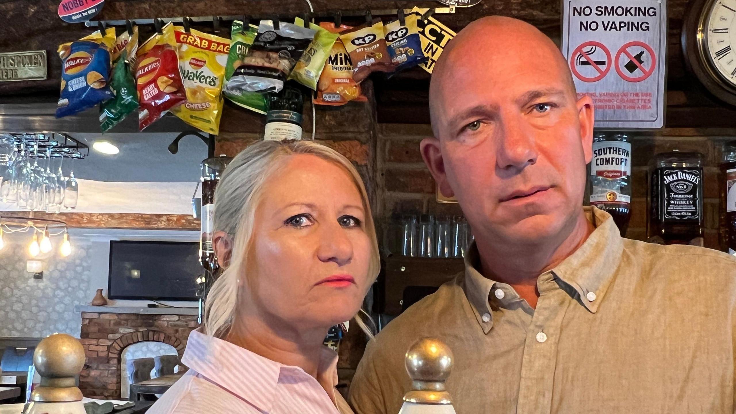 Julie and Lee Loach have stern looks on their faces as they stand behind the bar at their pub. Julie is wearing a striped pink blouse and has blonde hair. Lee is wearing a paisley brown shirt. Behind them, spirit bottles are fixed to the wall and crisp packets are strung above them.