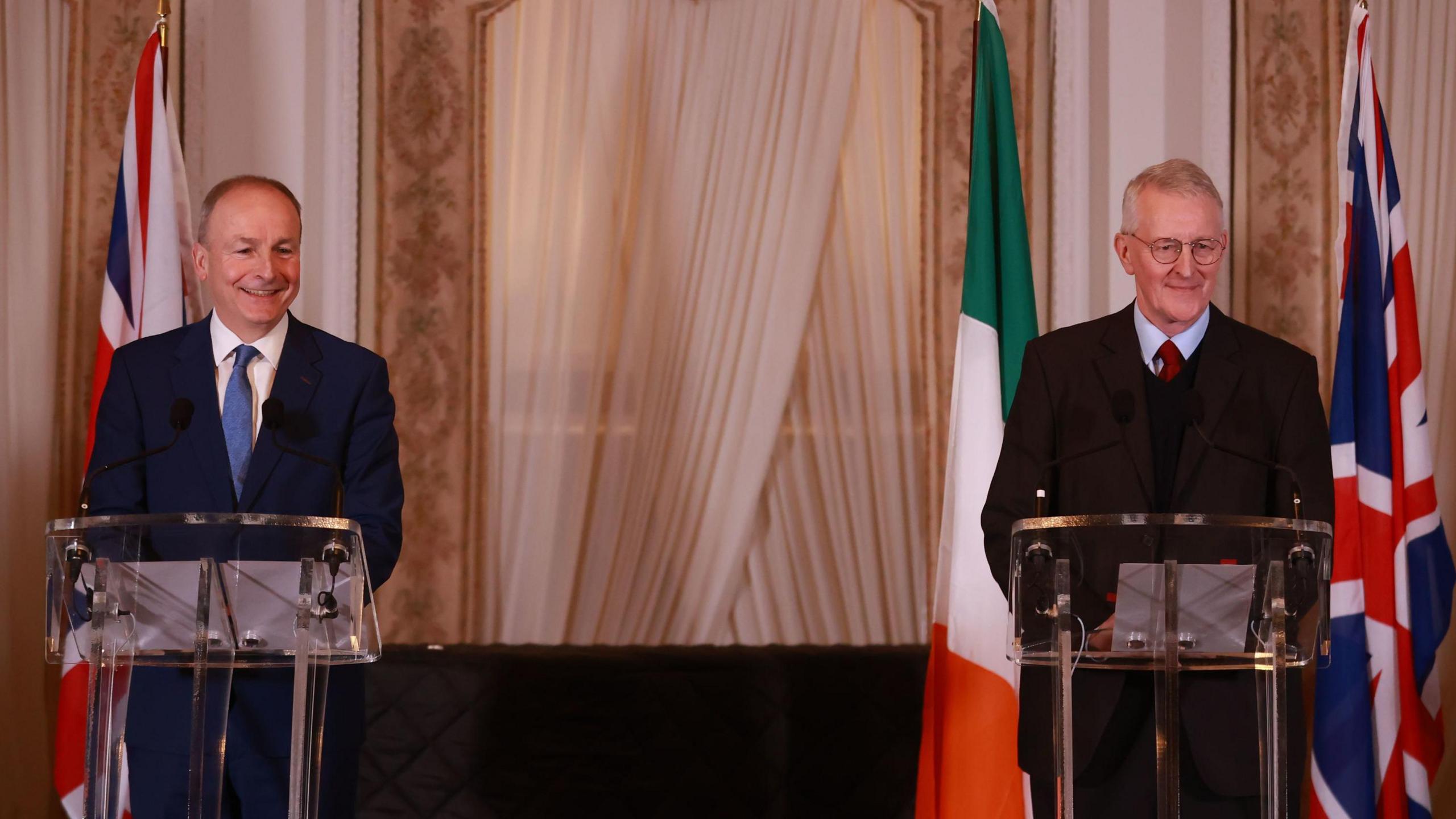 Micheál Martin and Hilary Benn stand behind transparent podiums. Martin is wearing a dark blue suit, white shirt and blue tie. Benn is wearing a dark suit, dark sweater, light blue tie and red shirt. Behind them are furled Irish and UK flags