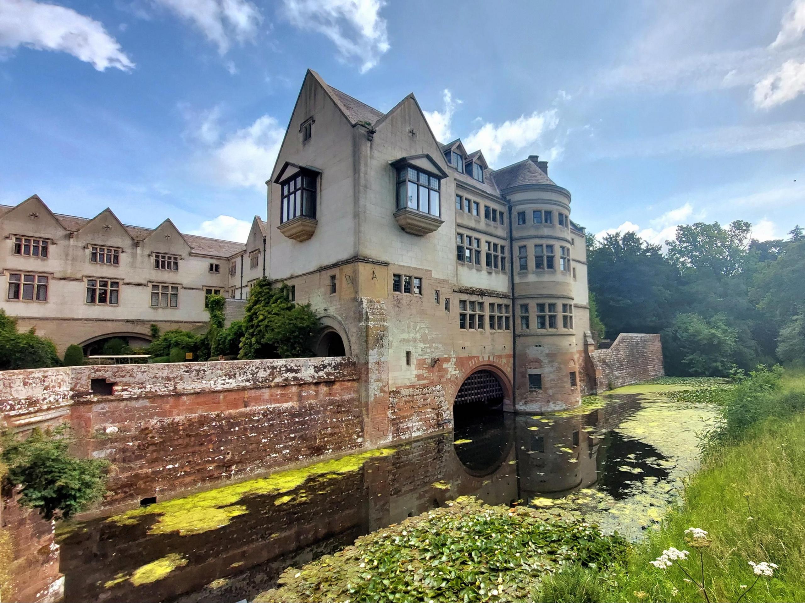 A grass field on the far right of the shot gives way to a moat, then a red stone wall with a honey-coloured turreted building overlooking the water, and behind it a longer block of buildings. 