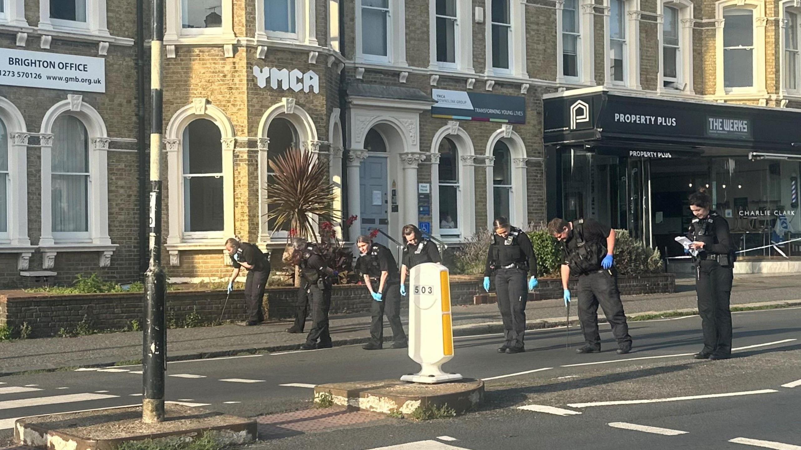 A line of police officers stood in the road. They are crouched over while searching the pavement and road surface.