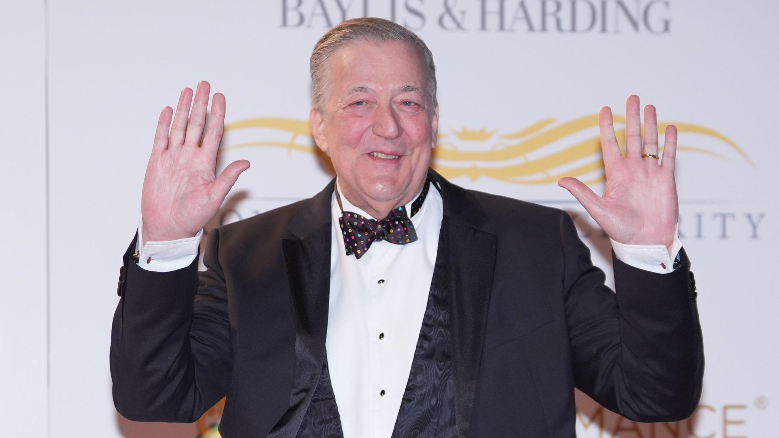 Sir Stephen Fry in a dinner jacket with a colourful spotty bow tie and his hands in the air