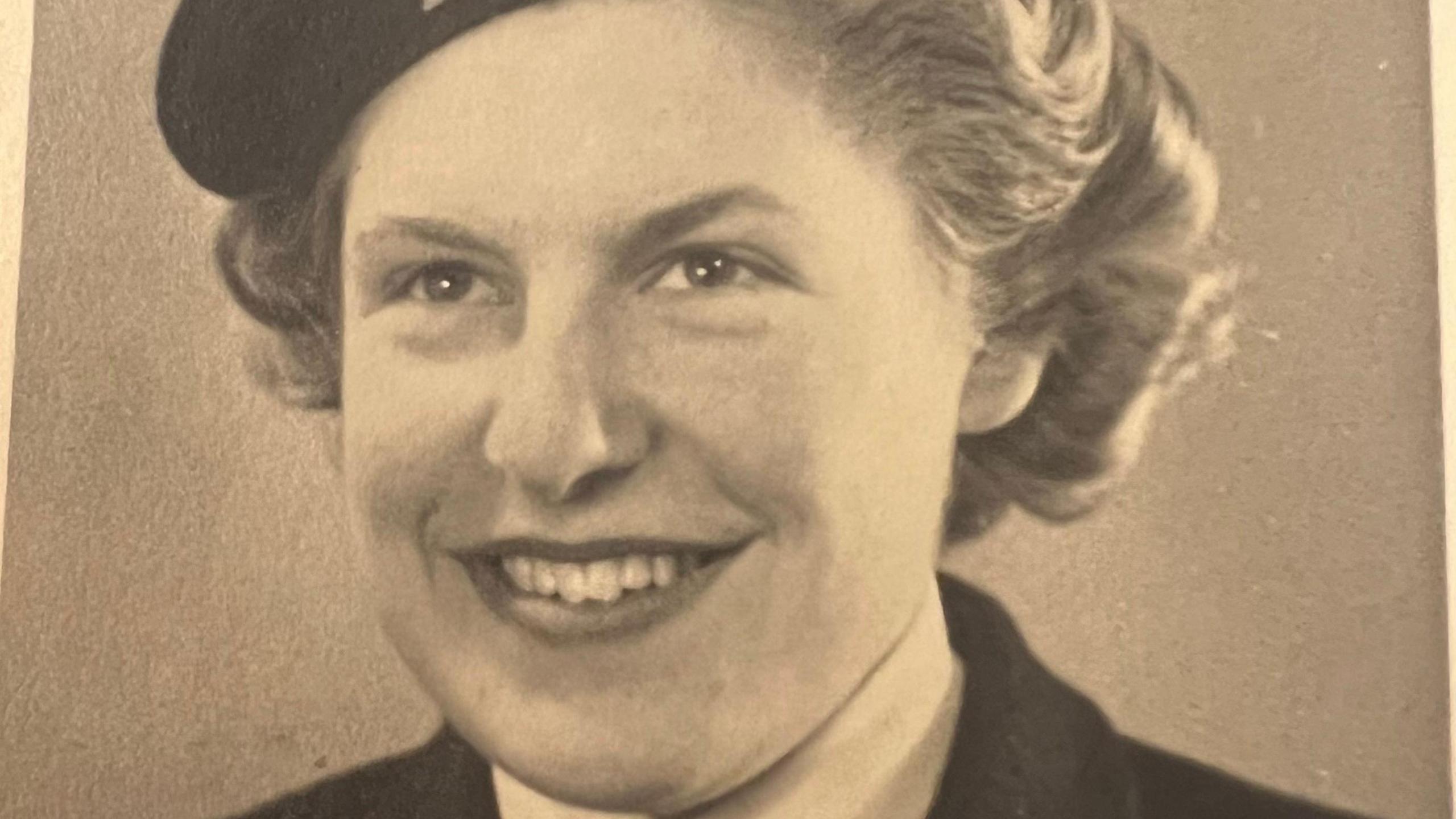 A sepia picture of a smiling woman with short hear and a dark jacket with a glimpse of a hat.