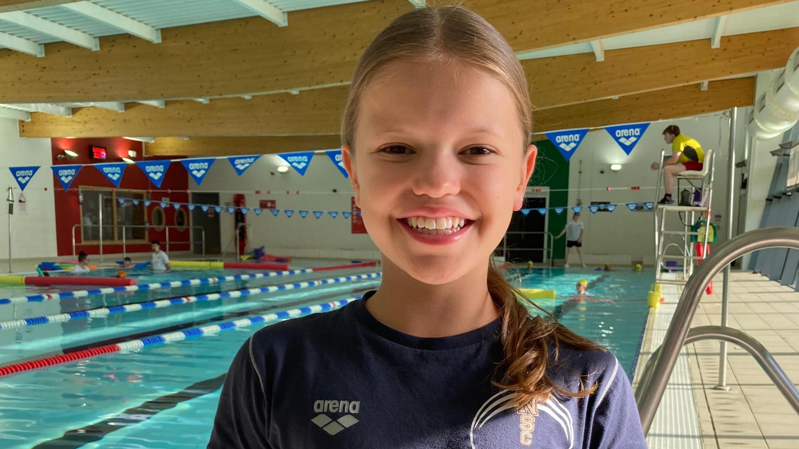 A girl with dark blonde hair in a ponytail pulled over one shoulder, wearing a blue T-shirt and standing in front of a bright blue swimming pool with a life guard on a tall chair to her right and some swimmers to her left