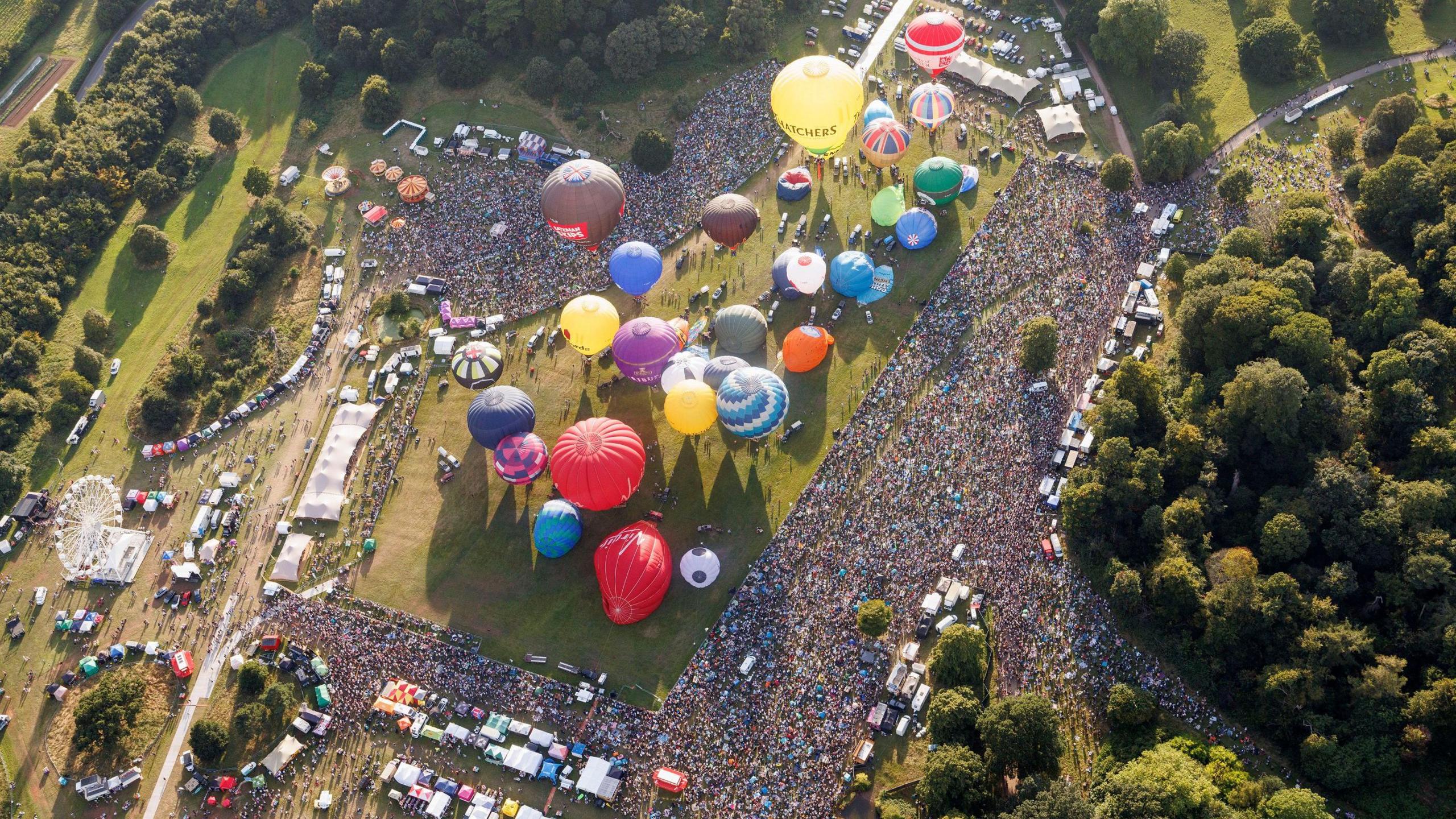 The Bristol International Balloon Fiesta 2024 site is seen from above. Thousands of people can be seen surrounding the main arena, where multiple balloons are inflated ready to take off. Stalls, other tents and vehicles can also be seen around the edge of the site