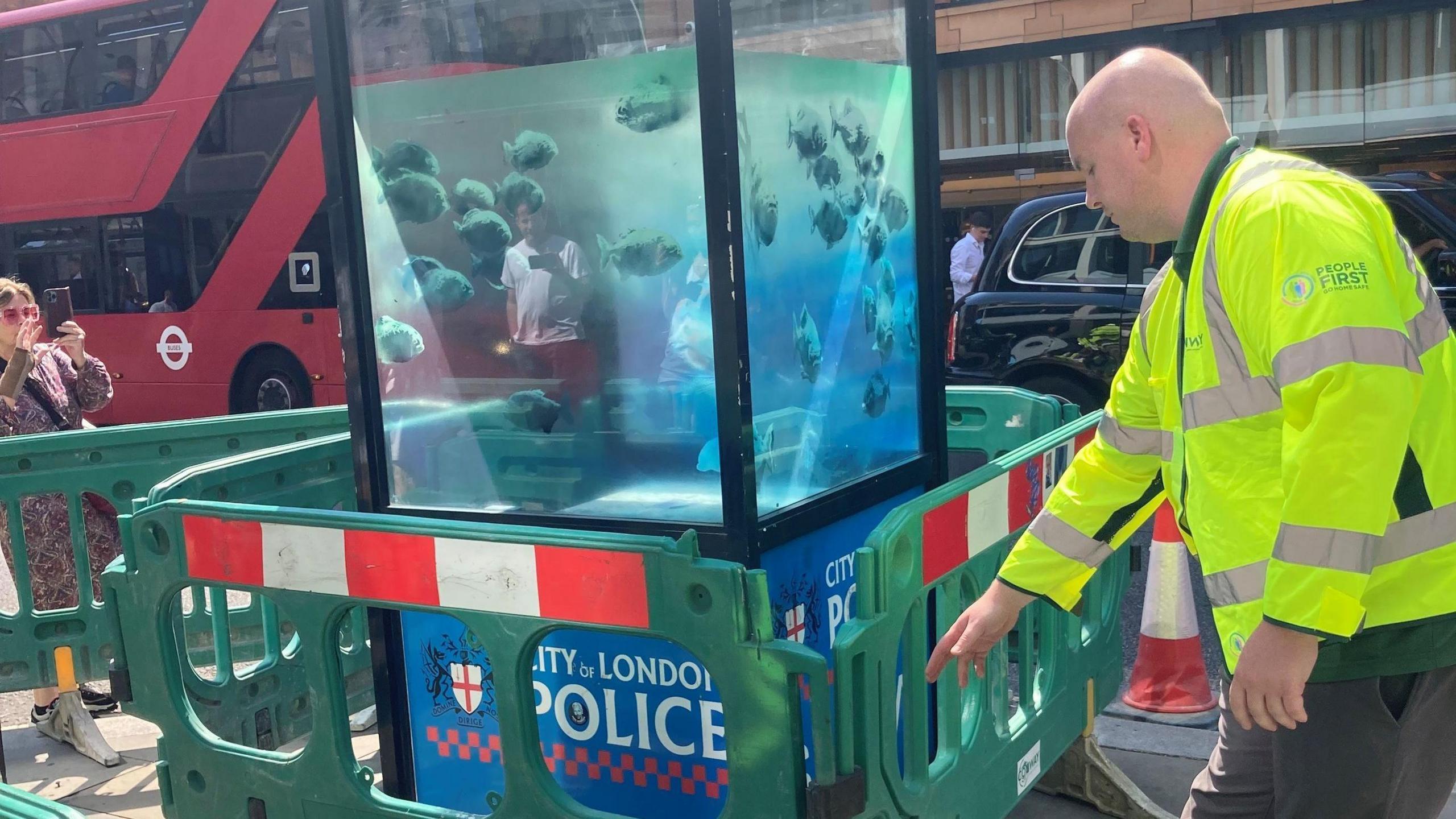 A City of London police sentry box painted in semi-opaque blue with piranhas in water, next to green, red and white barriers and a workman in yellow fluorescent clothing