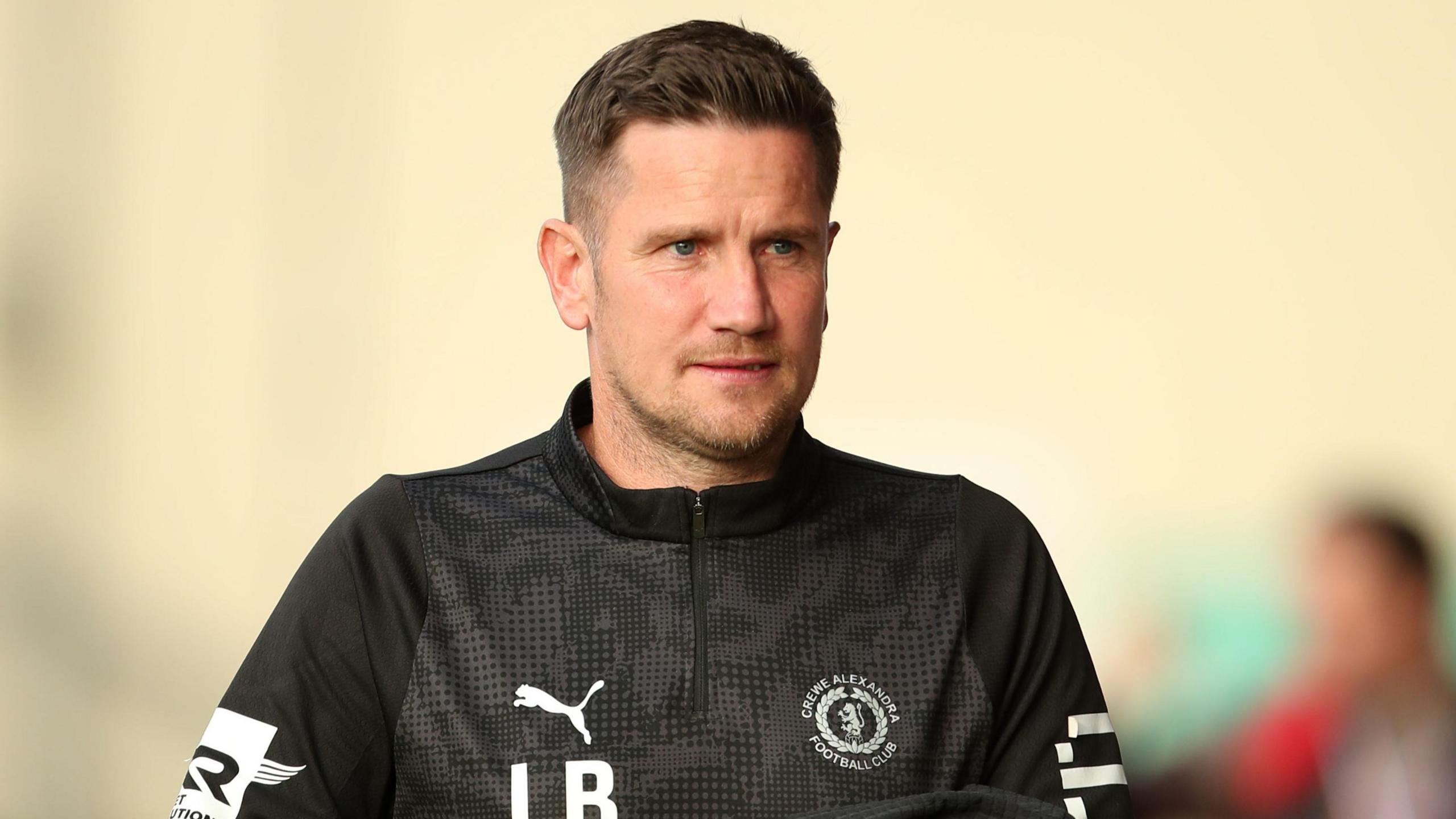 Crewe manager Lee Bell is about to open a bottle of water as he walks down the touchline in his black club track top