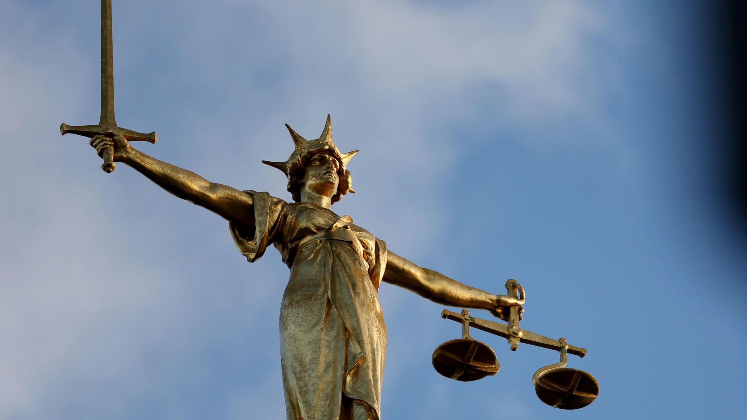 A close up of the statue of justice on top of the Old Bailey court building, a classical female figure rendered with a sword held aloft in one hand and a set of scales in the other hand.