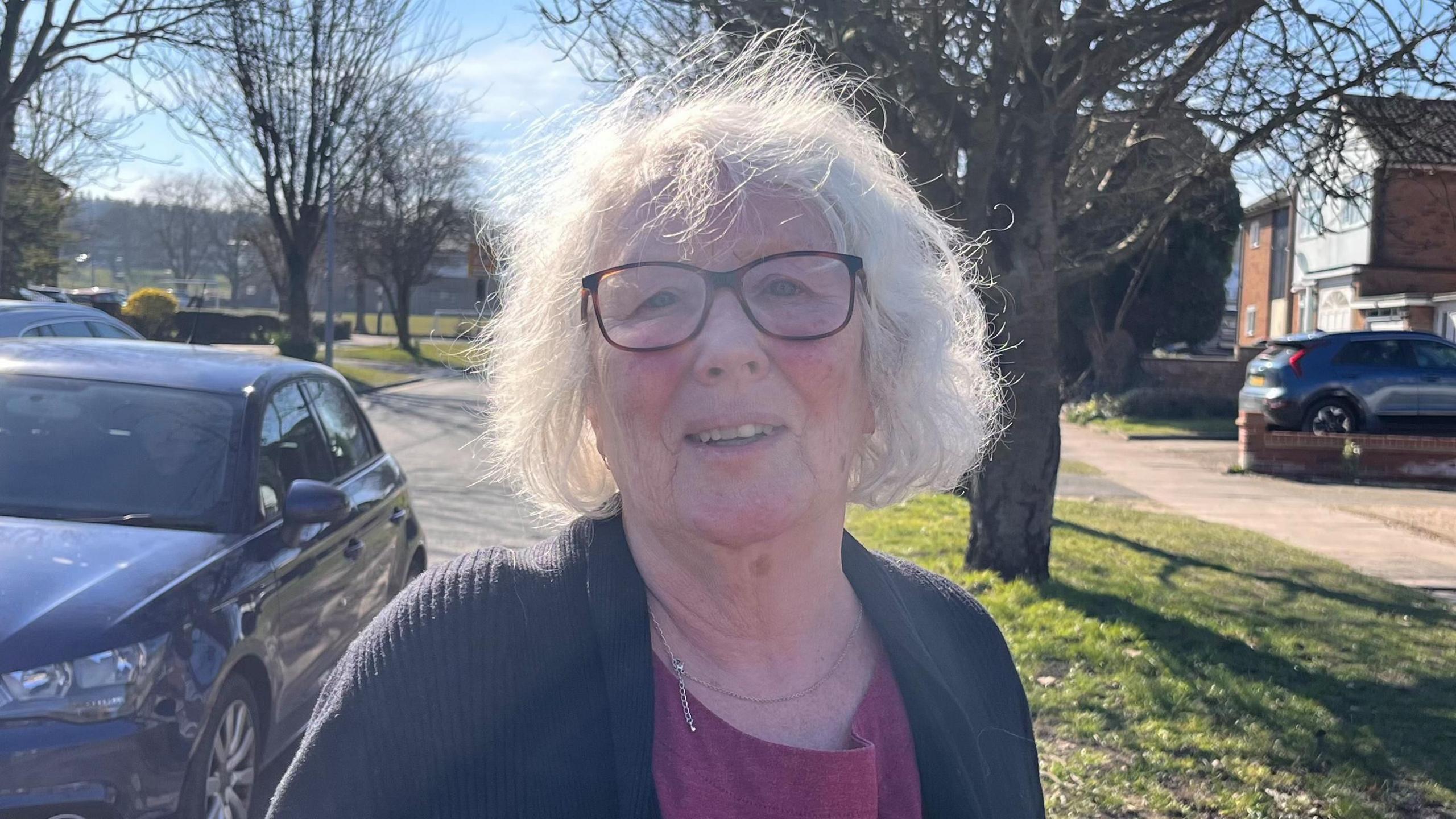 A woman with grey hair wearing glasses and a black cardigan and red top standing by a tree lined road with a car in the background. 