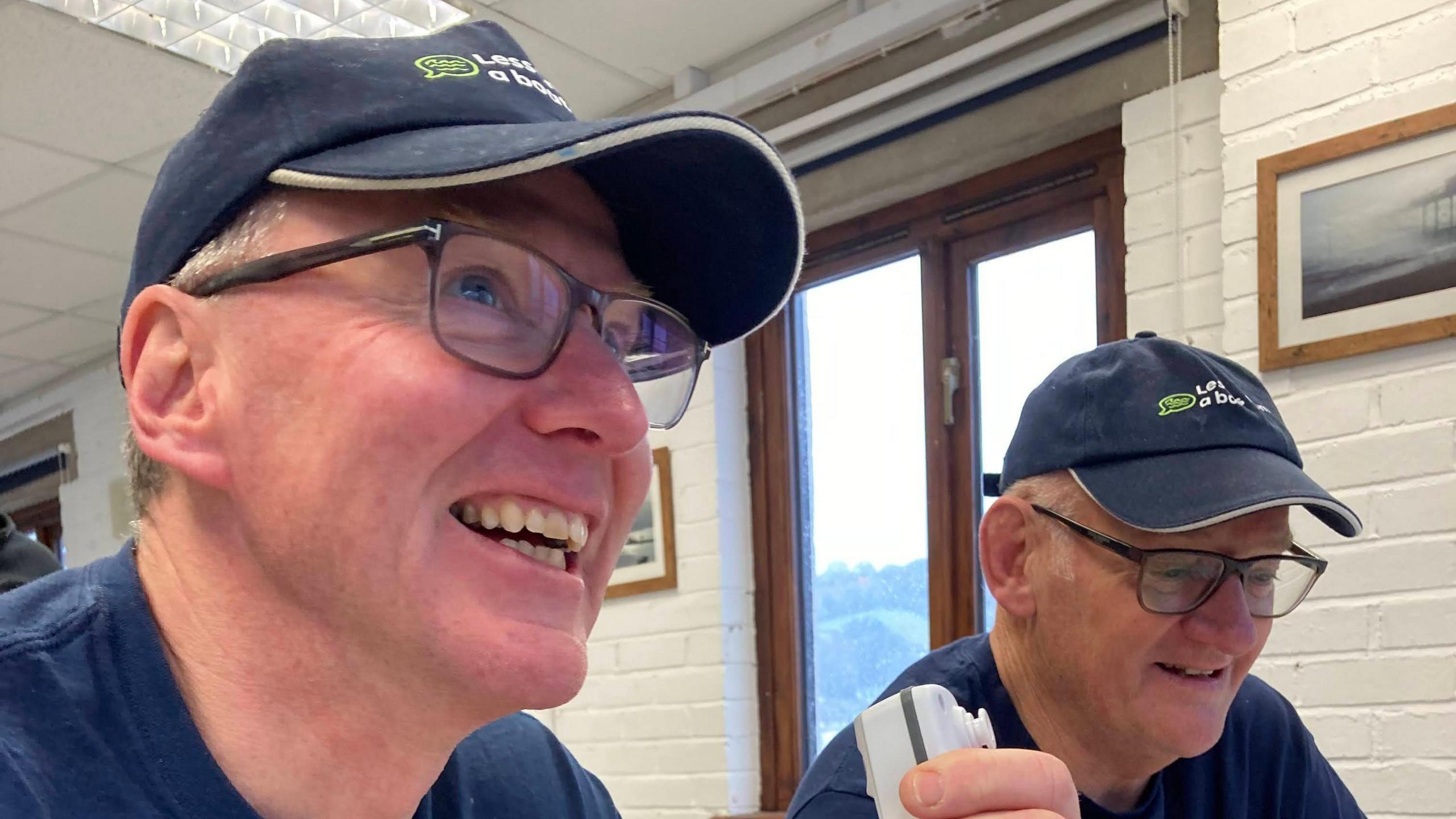 Two men in glasses, dark blue caps and T shirts smile while sitting at a table.