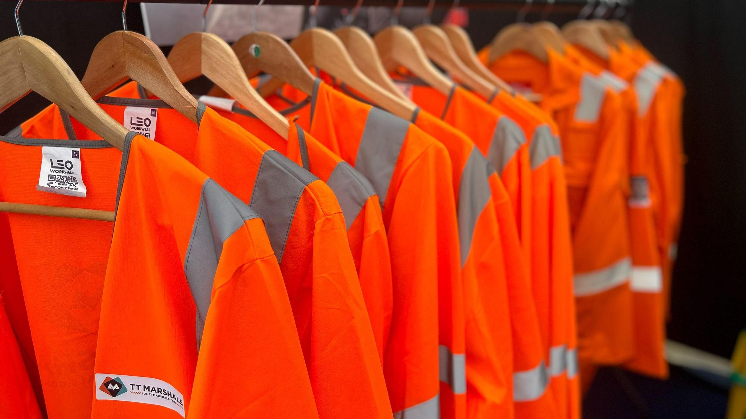 More than a dozen orange high visibility jackets hang on hangers.