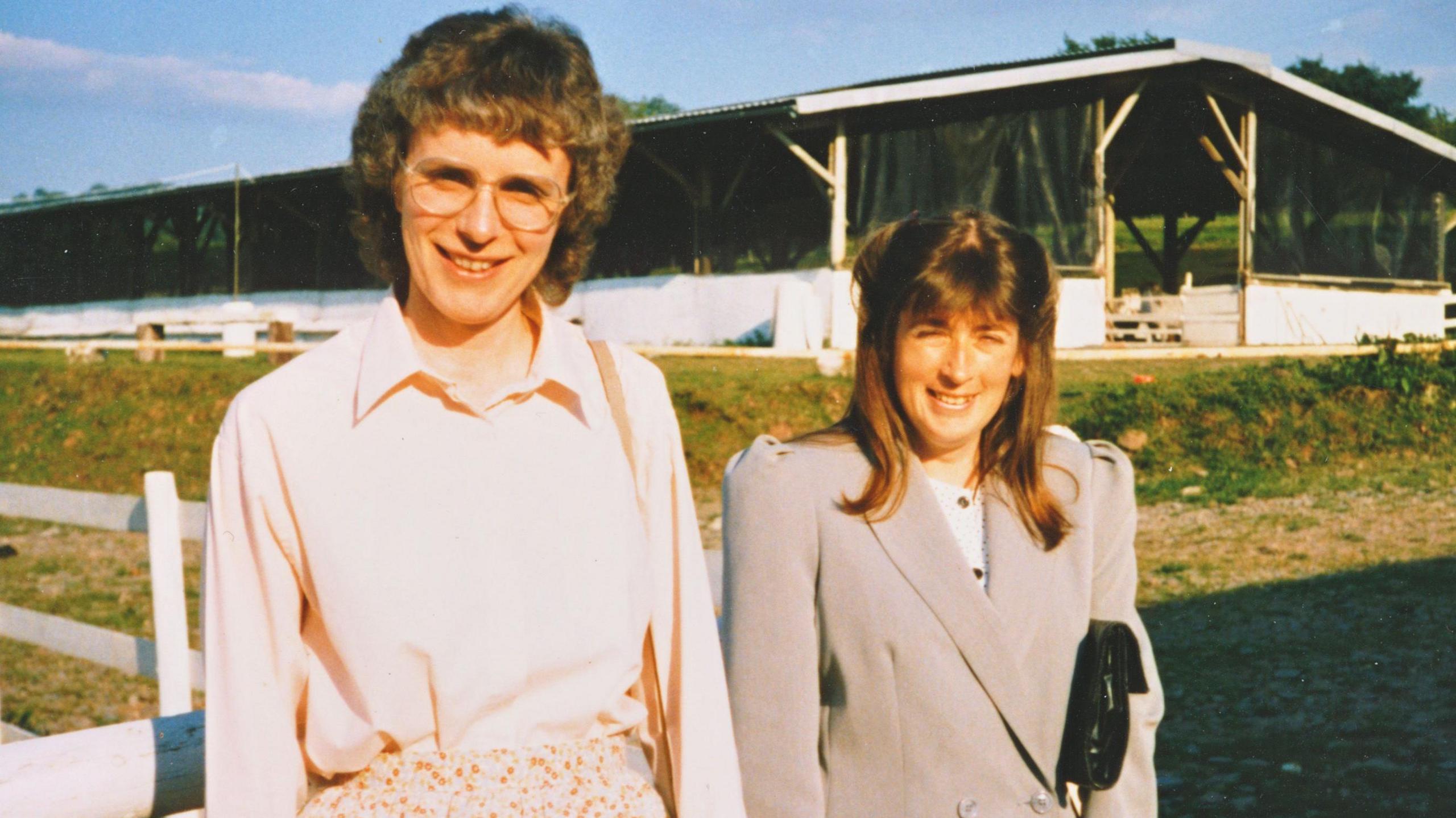 Barbara and Ginny at a farm. Ginny is wearing a beige blazer and has long brown hair with a fringe. Barbara is wearing a cream blouse and has short curly brown hair and glasses. 