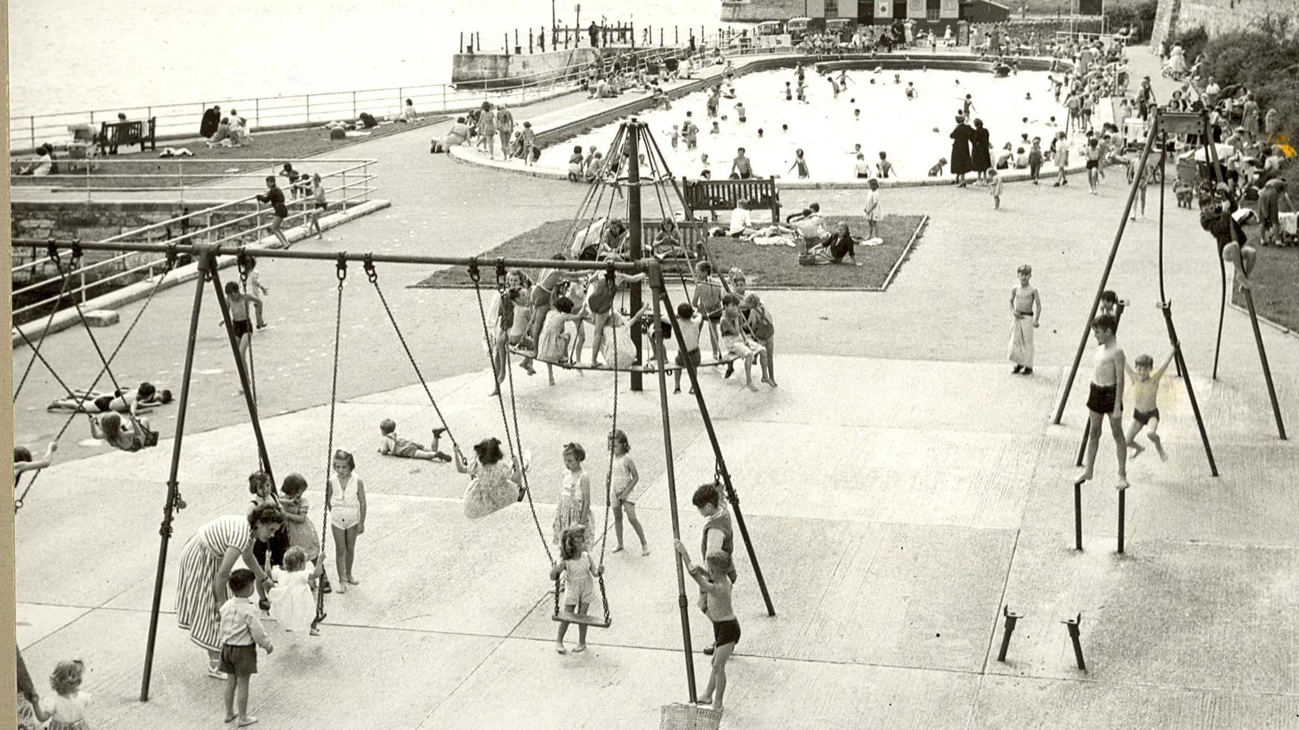 A black and white image shows children playing on swings with a swimming pool in the background at Mutton Cove.