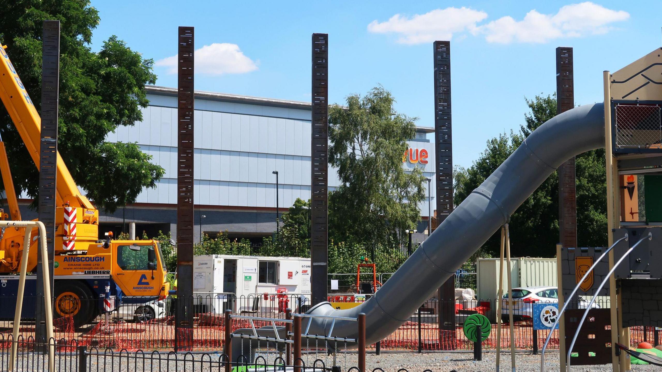 A park area being built, showing a large slide, large vehicle to the left, five heritage poles in place, and play equipment. A cinema building, that is large, and a grey box, is behind the building work.