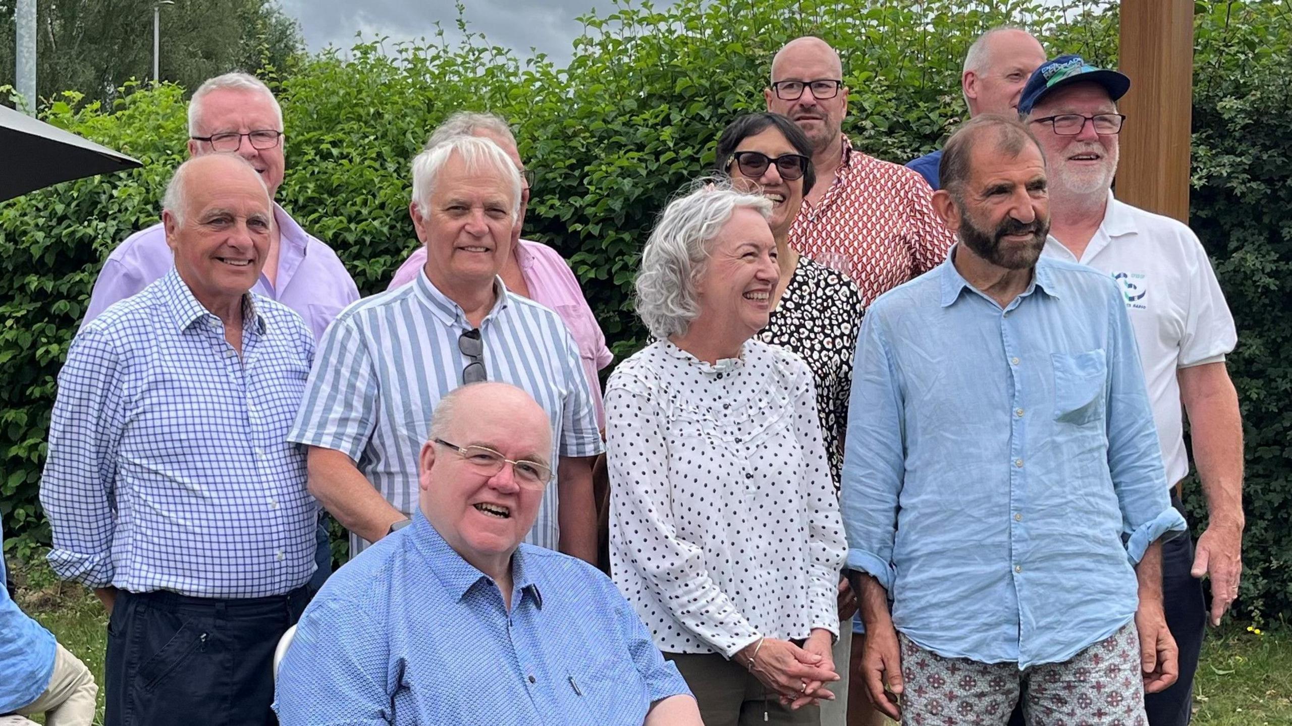 Nine men, all wearing either shirts or polo shirts, and two women, wearing dresses, pose outside for a picture. They are all smiling while standing on lawn in front of a bush.