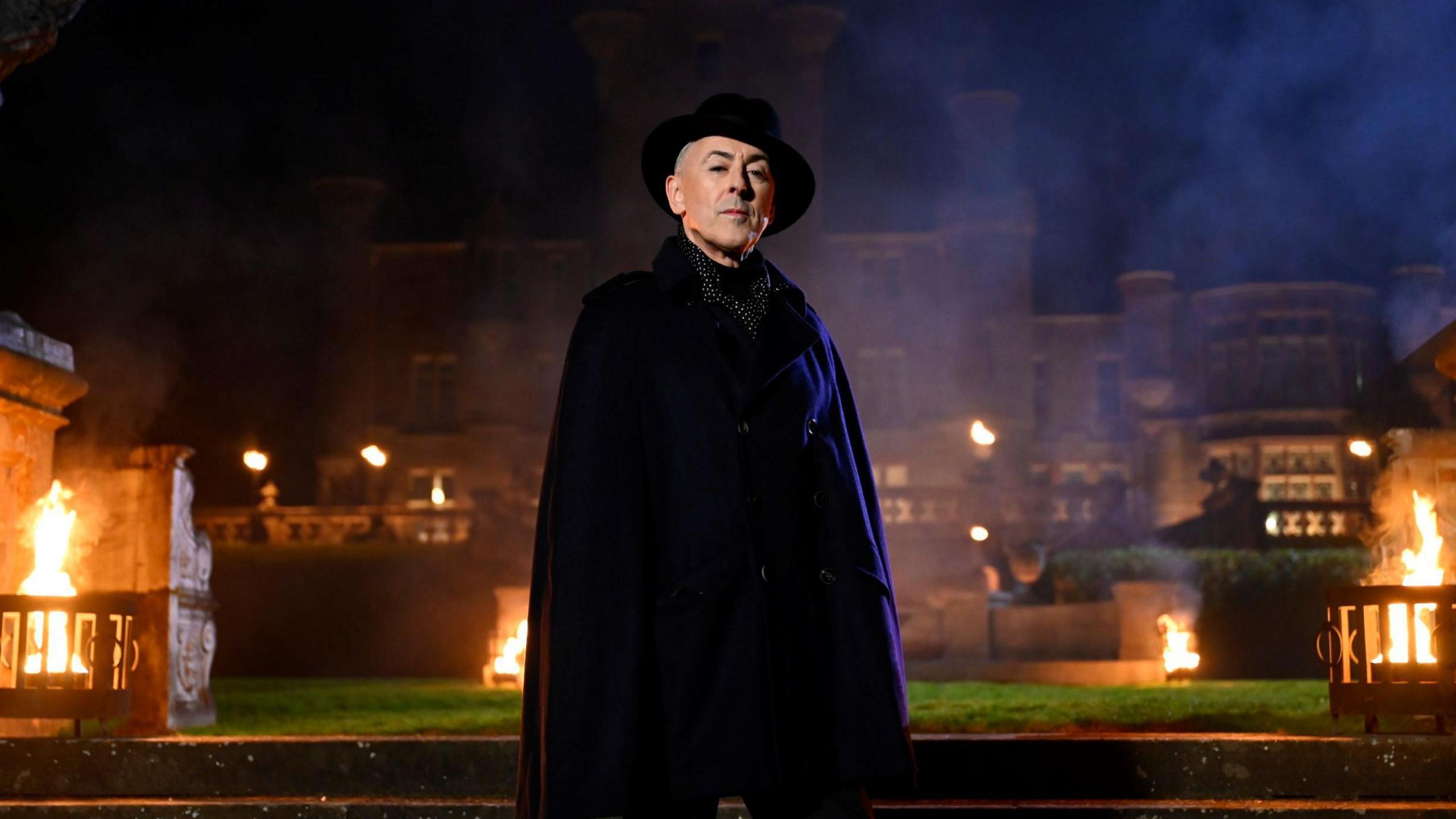 Alan Cumming standing in front of a castle at night, with various fires lit.  He is wearing either all black or purple, with a wide brimmed hat.  