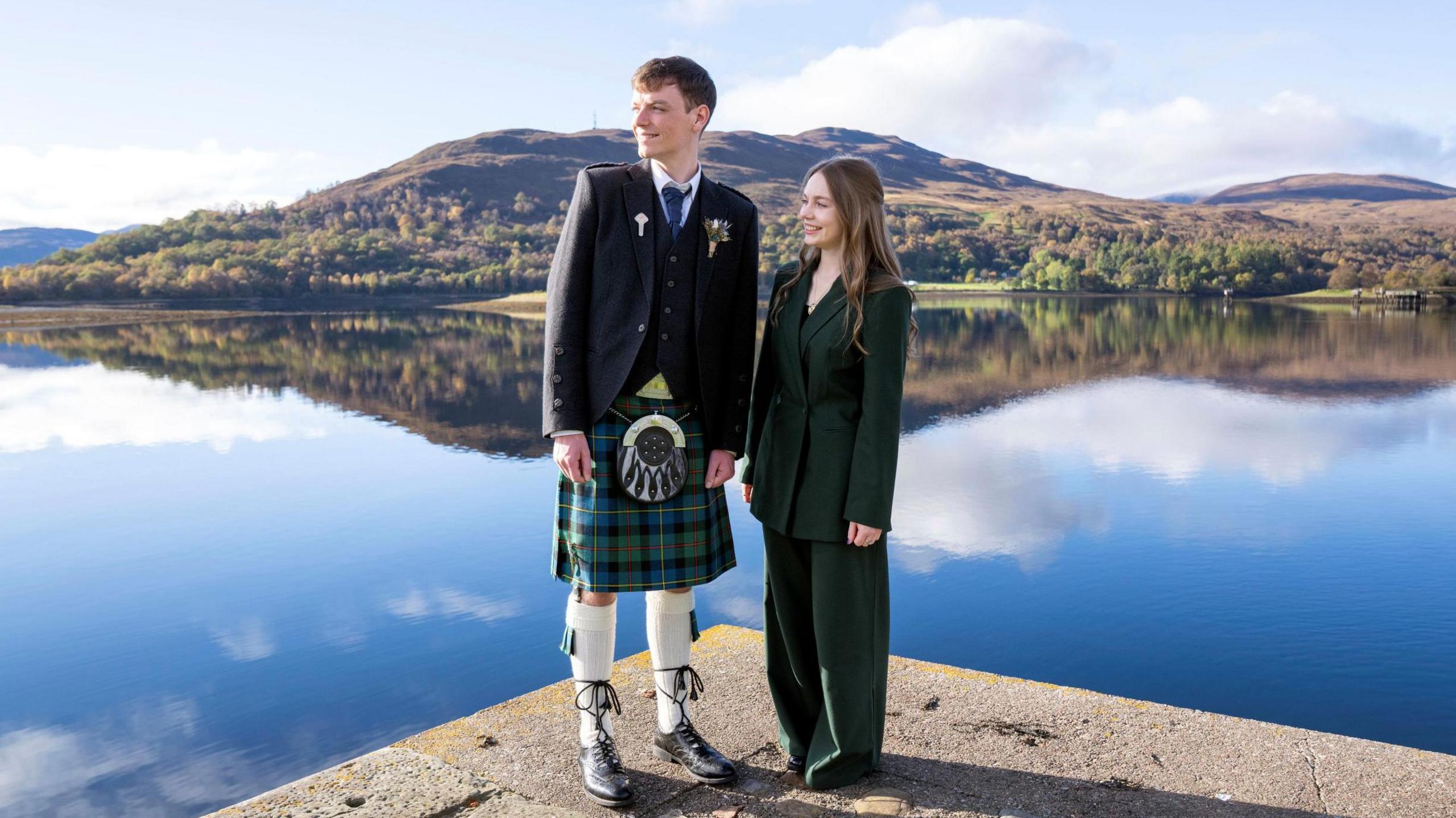 Duncan MacLeod and Mairi Callan are pictured outdoors in sunshine on a slipway on a loch. Duncan is wearing a kilt and jacket and Mairi a green trouser suit. The loch is flat calm and reflects the hills and woods on the opposite shore.