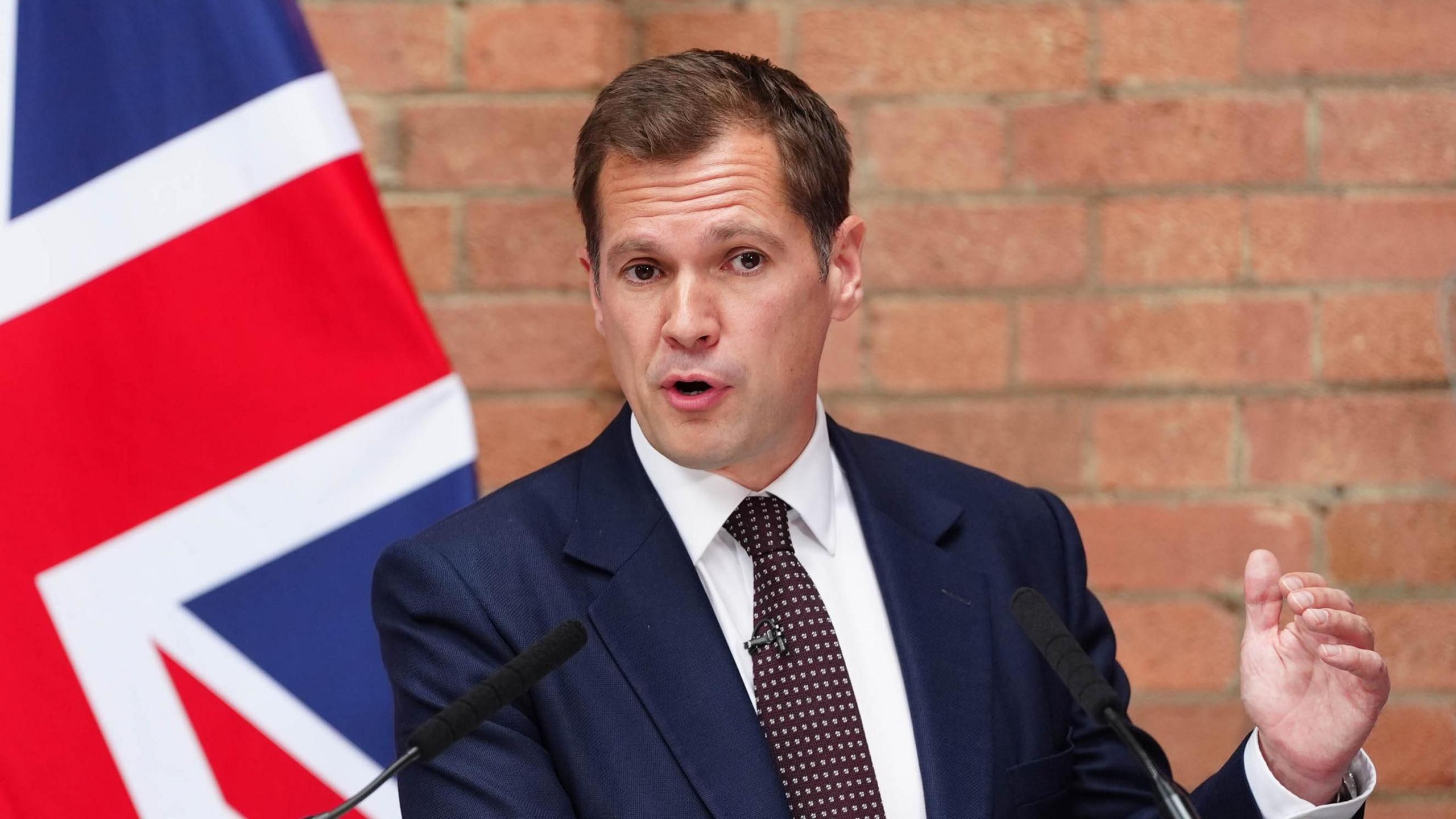 Robert Jenrick, in a suit and tie, makes a point at a press conference, with a Union flag and a brick wall behind him
