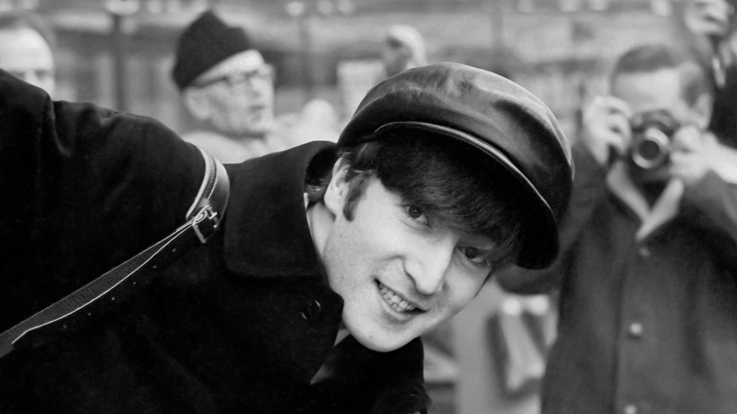 Titled: John on the Champs-Élysées, Paris, 15 January 1964. John wears a bakerboy-style hat, and a dark coat. He leans into the camera frame and smiles. A photographer is seen, blurred in the background.