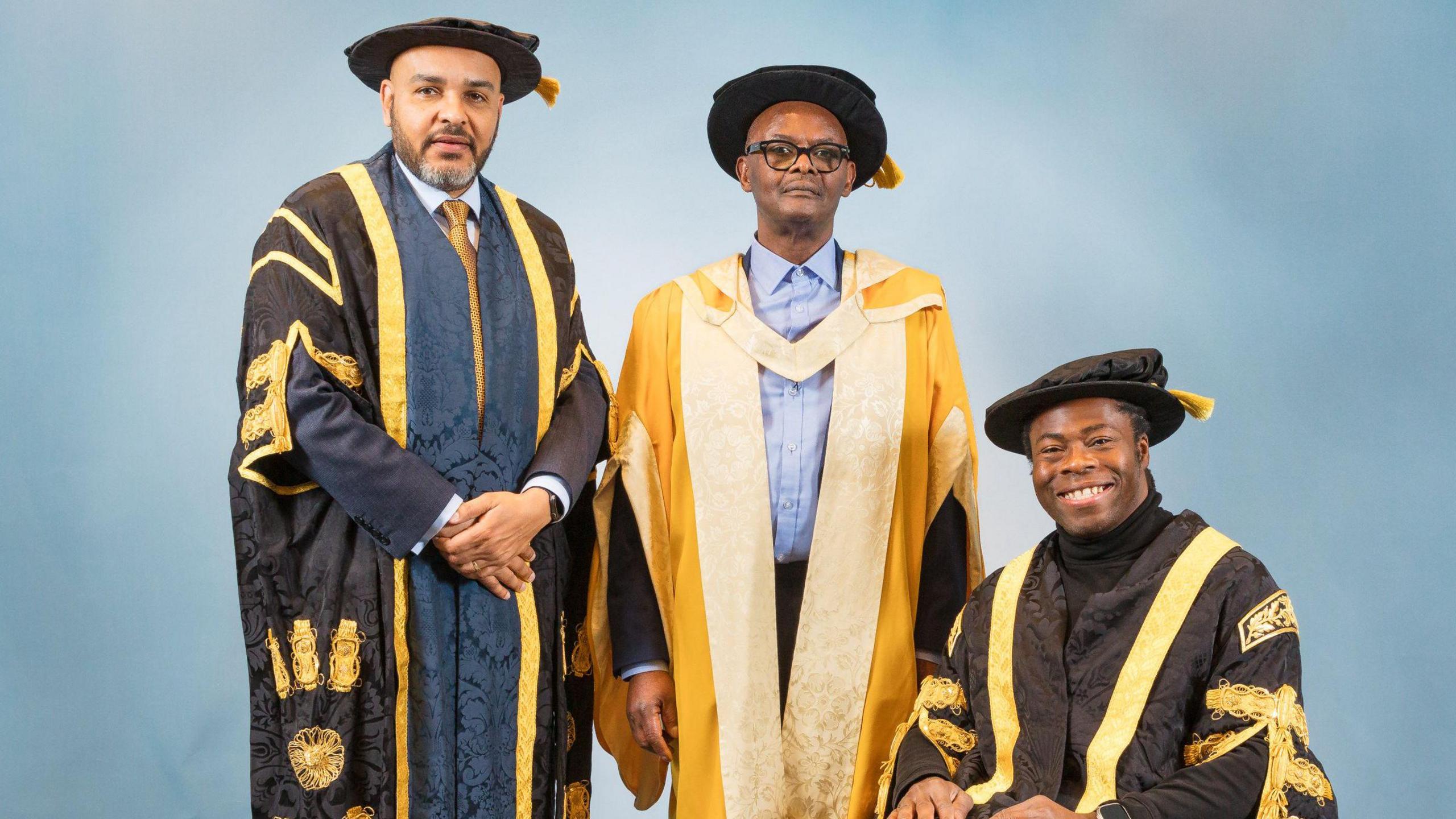 Dr Vanley Burke, dressed in a gold graduation gown stood in the middle of two men. One is standing and one is sitting in a wheelchair.