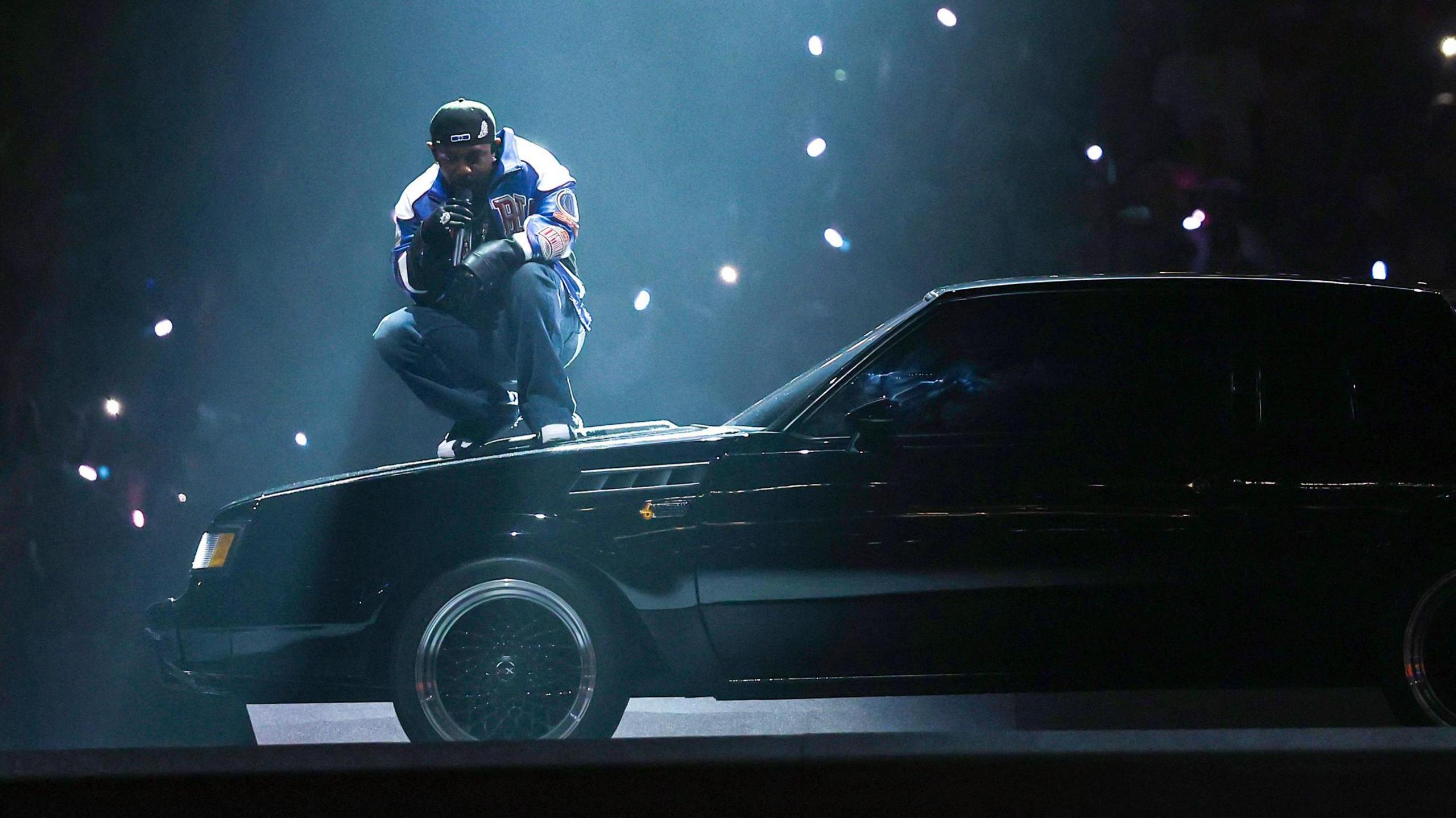 Kendrick Lamar during his halftime show at the Super Bowl. The stadium is dark with the rapper lit by a single spot light as he crouches on the bonnet of a black muscle car. 