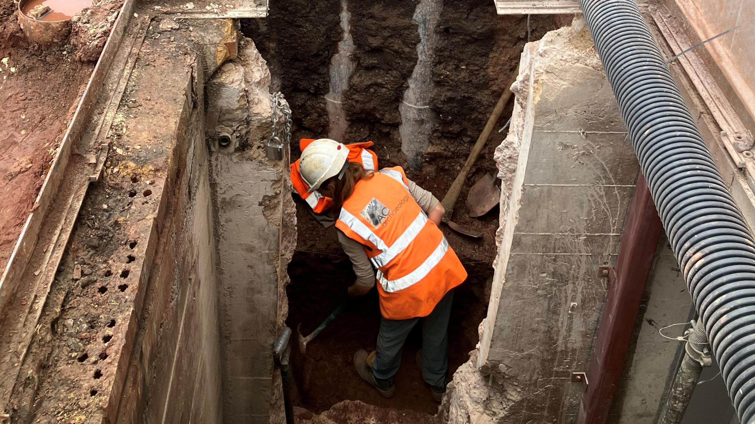 A member of the AC Archaeology team digging in a deep pit. They are wearing an orange high-vis jacket with a white hard hat.