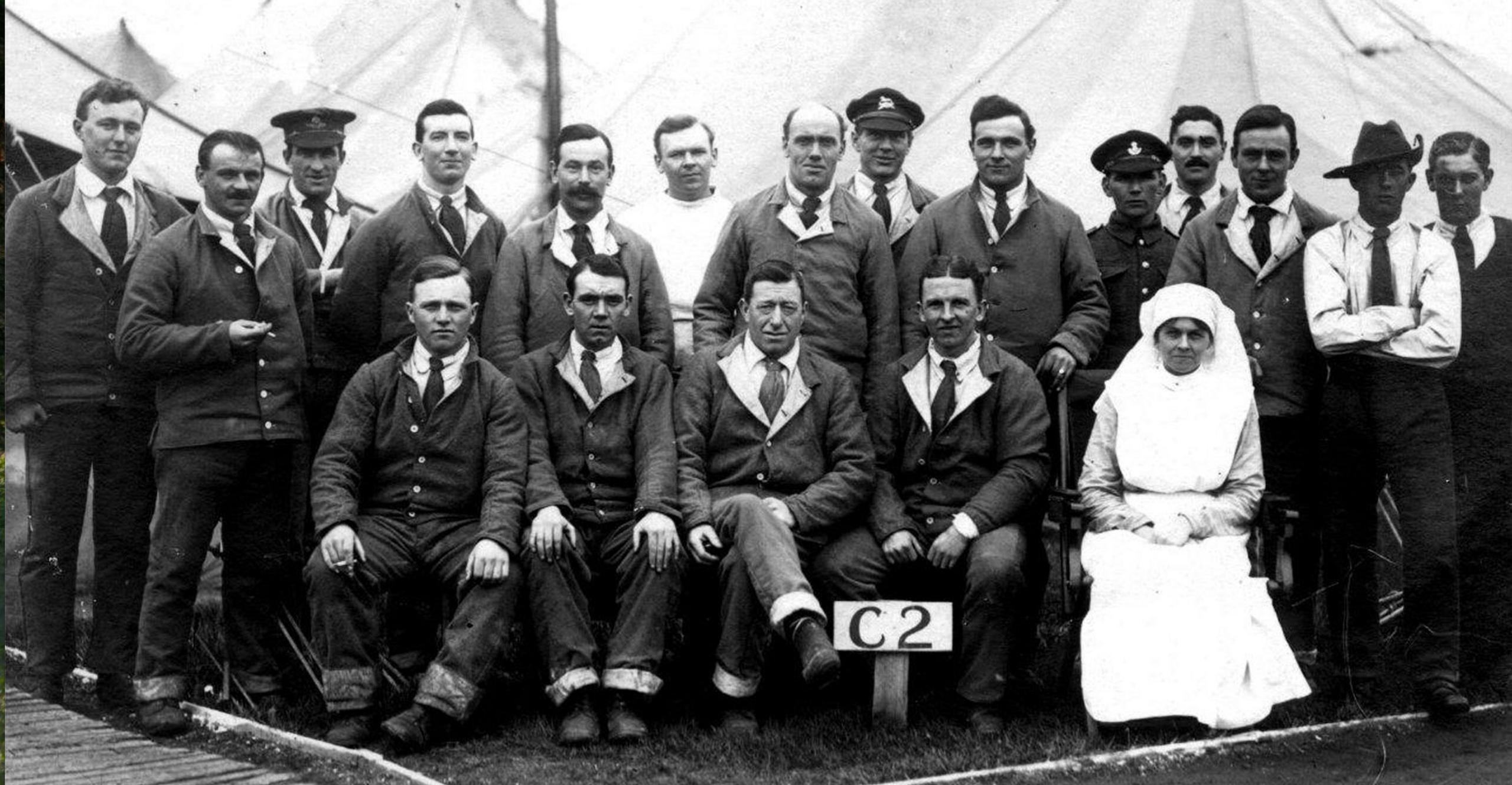 The black and white image shows a group of men, in cotton trousers and tops, sitting with a woman in a nursing uniform inside a tent. 'C2' is written on a sign. The photo, taken in 1916, is believed to be men from the 14th battalion, Worcestershire Regiment, recuperating at a field hospital.