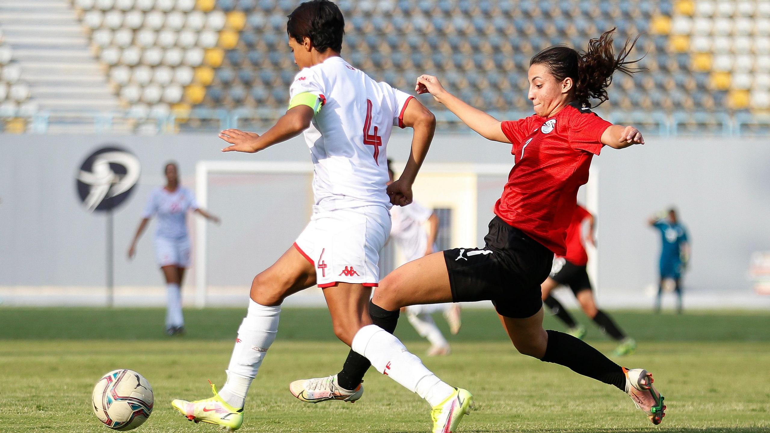 An action shot during a football match shows Nadine Ghazi, wearing a red Egypt shirt, black shorts, black socks and white boots, spreading her arms for balance and stretching her left leg forward as she challenges an opponent in an all-white strip for the ball. Four other players, a goal and an empty stand can all be seen out of focus in the background.