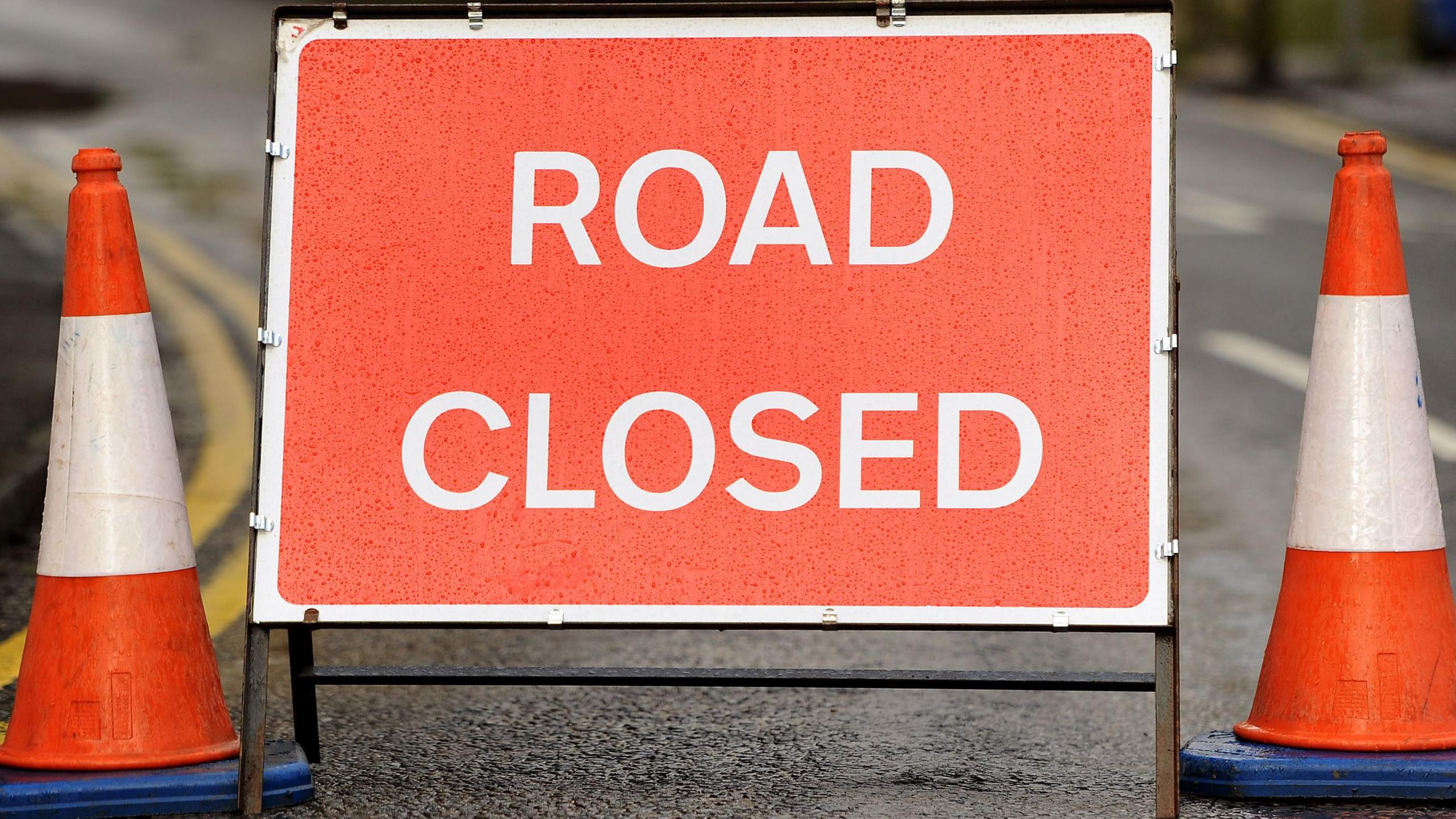 Close up of red road closed sign. Traffic cones are on either side of the sign.