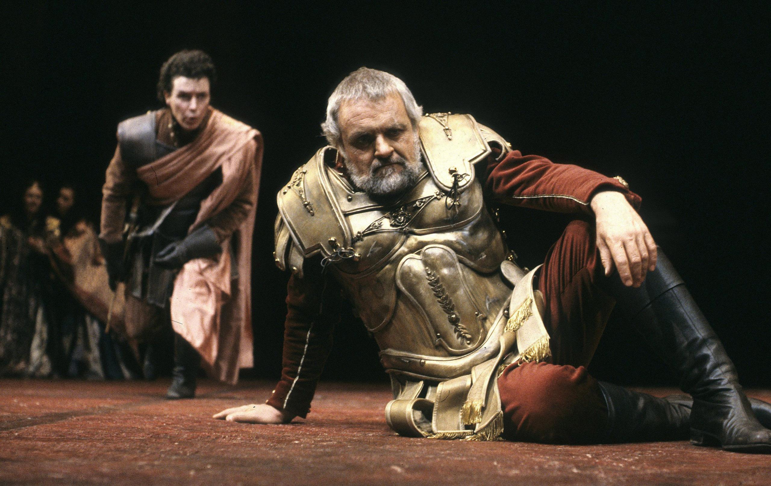 Sir Anthony on the ground in gold armour and black boots, behind him another actor crouching with pale pink sash wrapped around him in Anthony and Cleopatra at the National Theatre in 1987