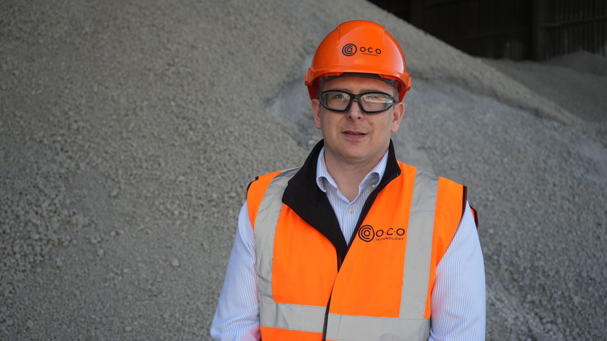 Graham Cooper in front of a pile of limestone aggregate. He wears an orange hard hat with O.C.O on it and an orange hi-vis vest over a blue and white pinstriped shirt. He has black rimmed goggles on.