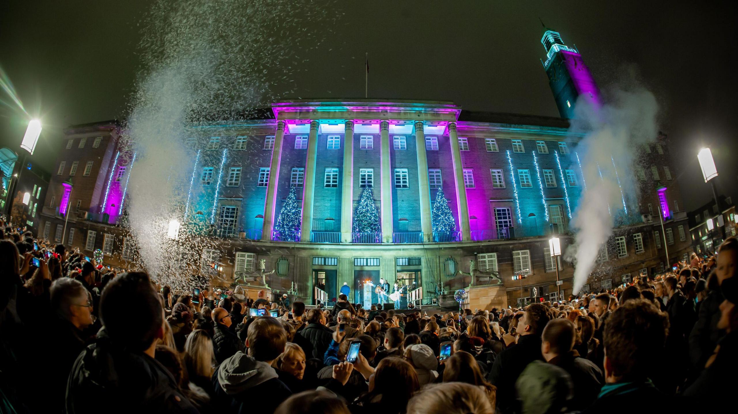 Norwich City Hall, with its numerous large columns, lit up in blue and purple. Christmas trees adorn the balcony and hundreds of people are stood outside, many of them holding up their mobile phones.