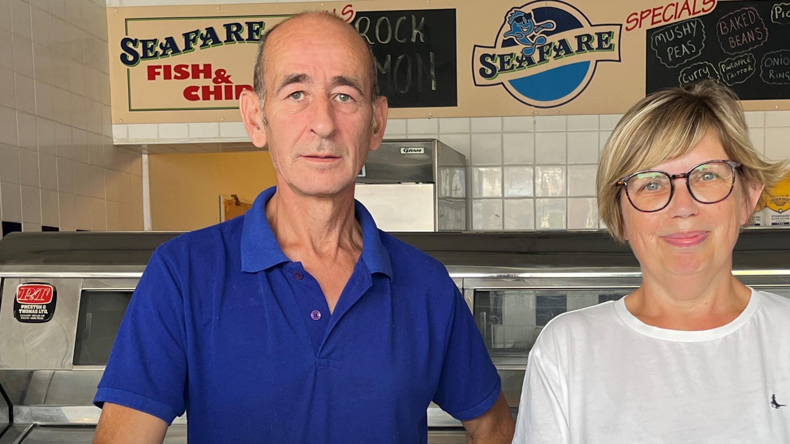 Iain Moore dressed in blue and Carol Moore in white stand in their fish and chip shop