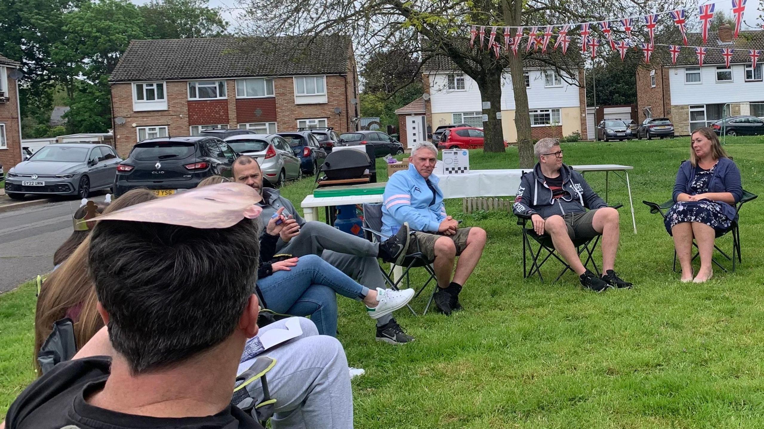 Neighbours sitting on camping chairs on Catherine Drive enjoying the green for a celebration. There are a number of cars and houses in the background.