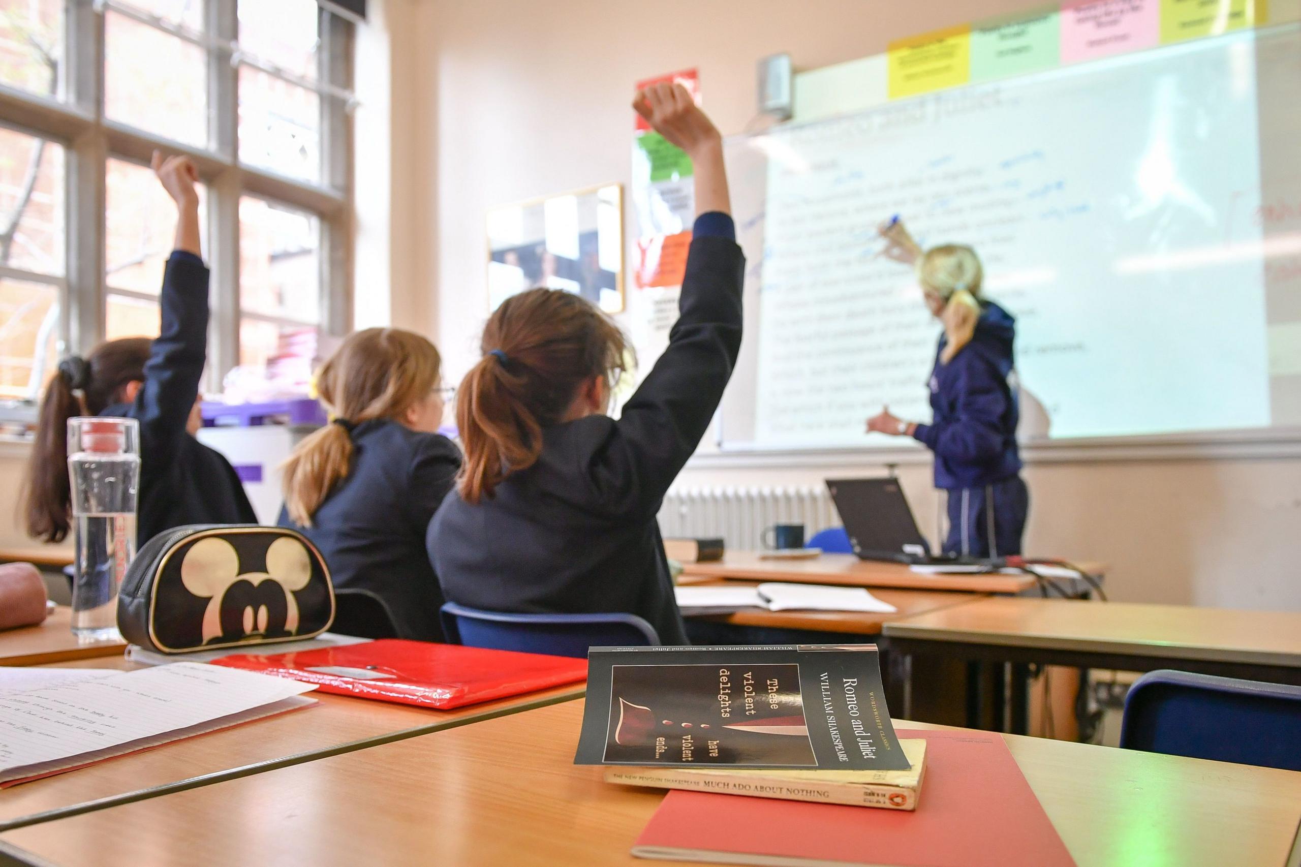 A generic photograph of a classroom, showing children raising their hands to a teacher's question