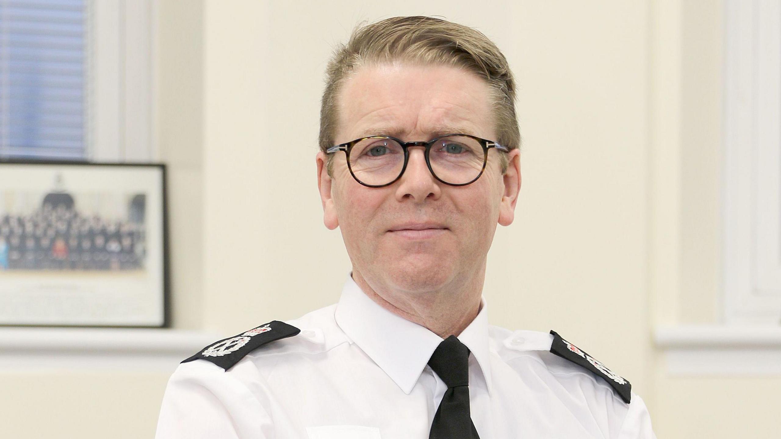 Chief Constable Will Kerr stands in front of a Devon and Cornwall Police sign. sits in an office. He is wearing a white shirt with a black tie and black epilates. A framed photo of police officers is behind him.