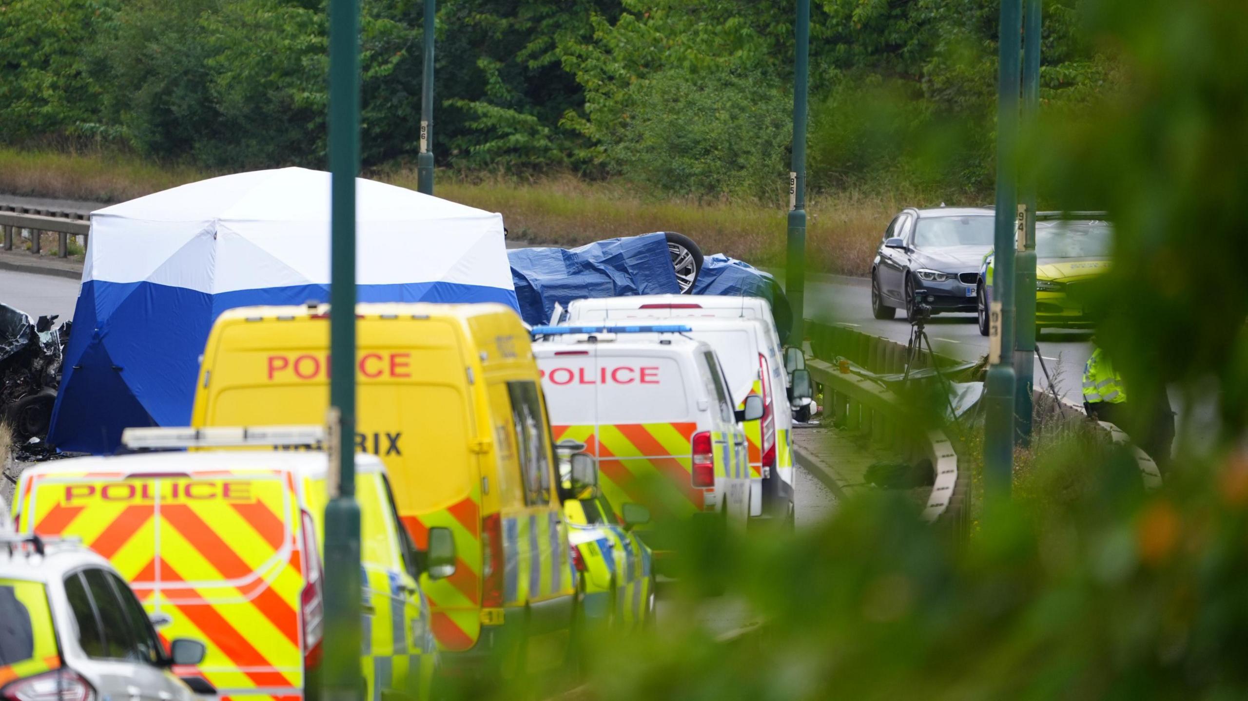 An overturned car is seen under a tarp, surrounded by police vehicles on a road.