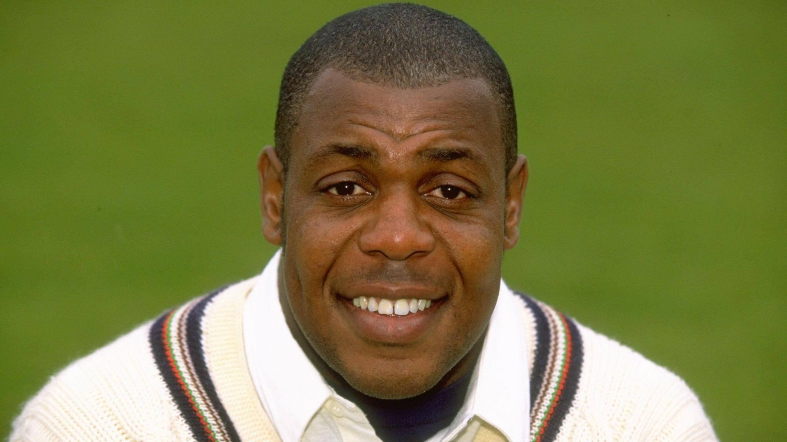 David 'Syd' Lawrence. He is has brown eyes and short dark hair. He is pictured wearing a white cricket jumper with a stripe around the neck. He is looking at the camera and smiling.