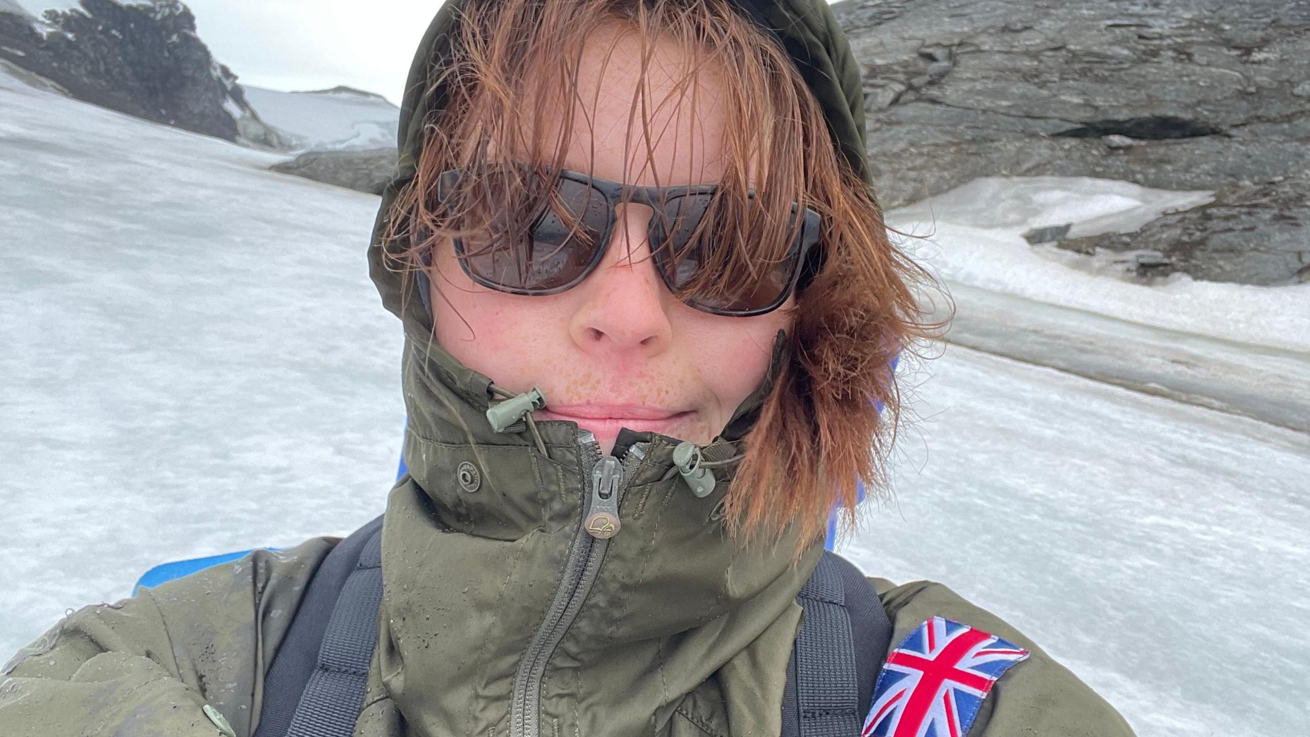A selfie of Emily Broadwell wearing a khaki raincoat and black sunglasses with her hood up, and her gingery-brown hair poking out. Snow and ice on rocky mountains surround her.