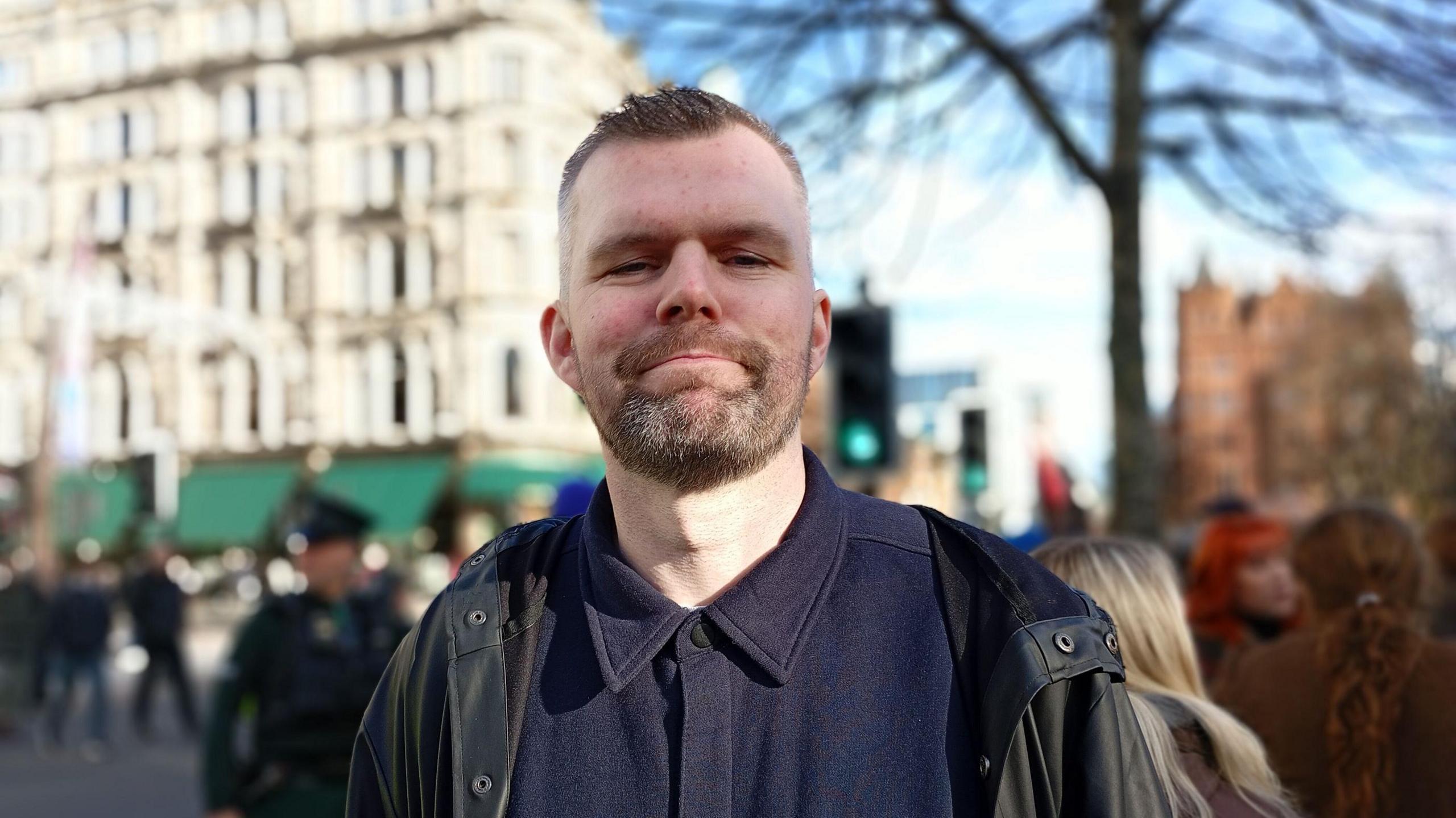 Gerry Carroll at the Givan Must Go rally in Belfast.   He has short, greying hair and a beard.  He is wearing a dark waterproof coat over a navy shirt.  There are tall buildings, a tree and traffic lights behind him.