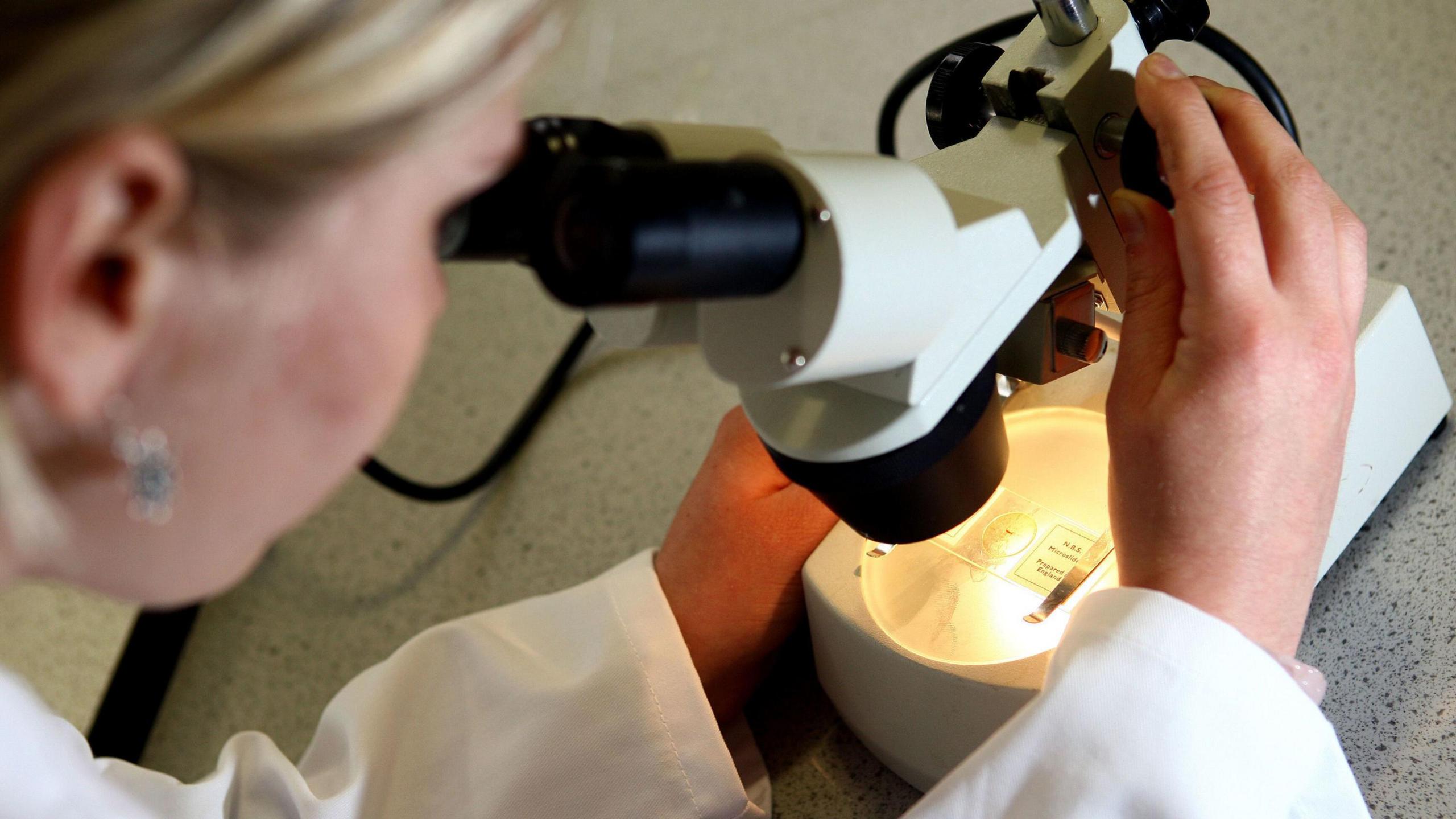A stock image of a woman dressed in a white lab coat looking through a microscope.