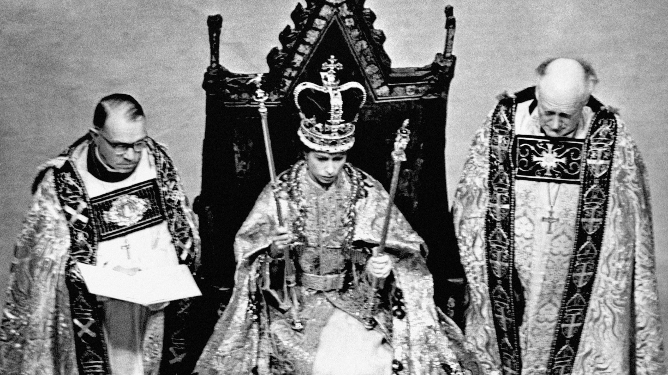 A black and white photo of Queen Elizabeth II wearing the St Edward's Crown at her coronation. She is holding two sceptres and flanked by two clergymen.  