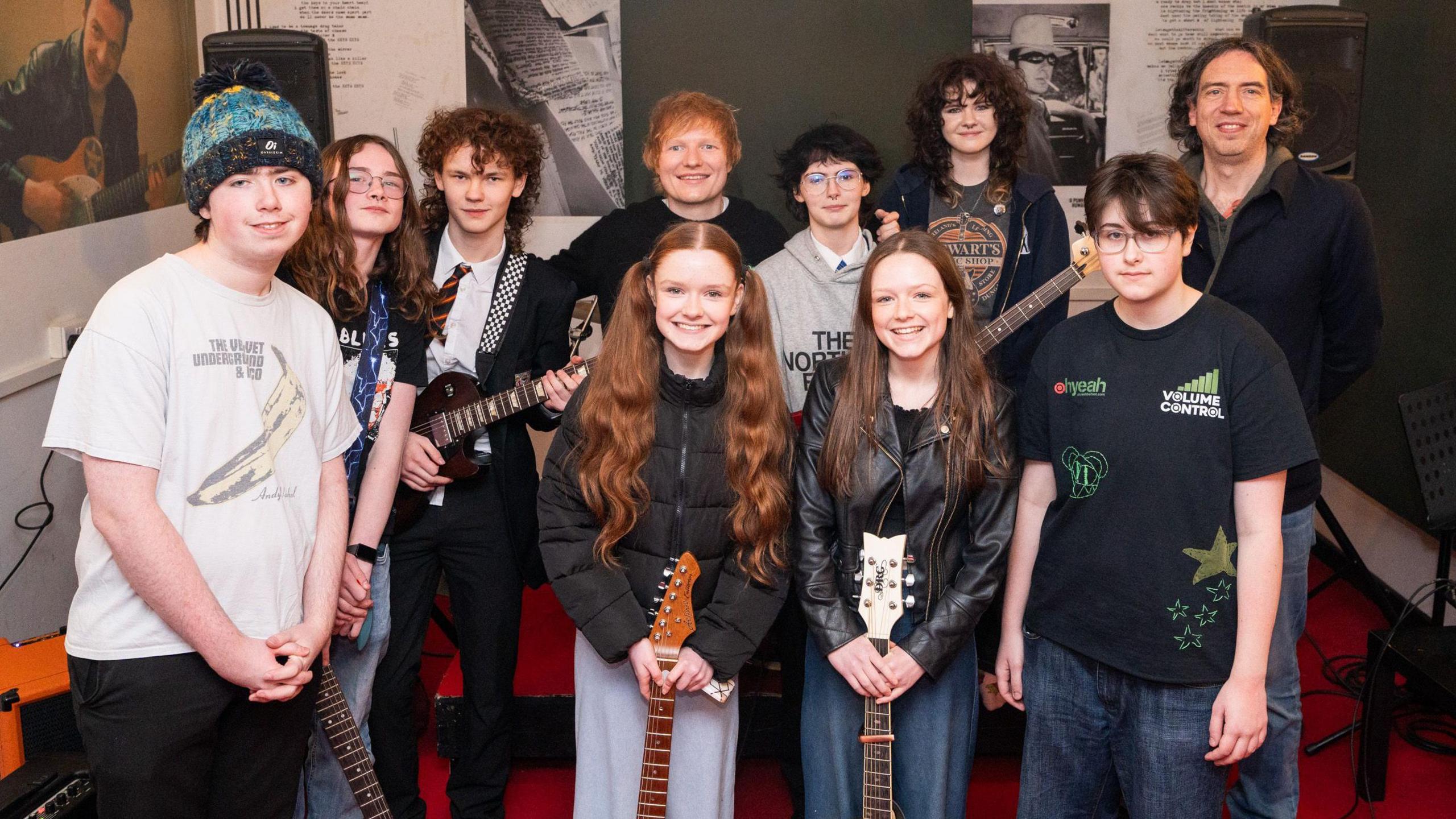 Ed Sheeran stands at the back of a large crowd and smiles while he poses with young people. On the right hand side is Gary Lightbody. Eight other young people in the room, including Ruby and Molly holding guitars in the front row, are smiling at the camera.