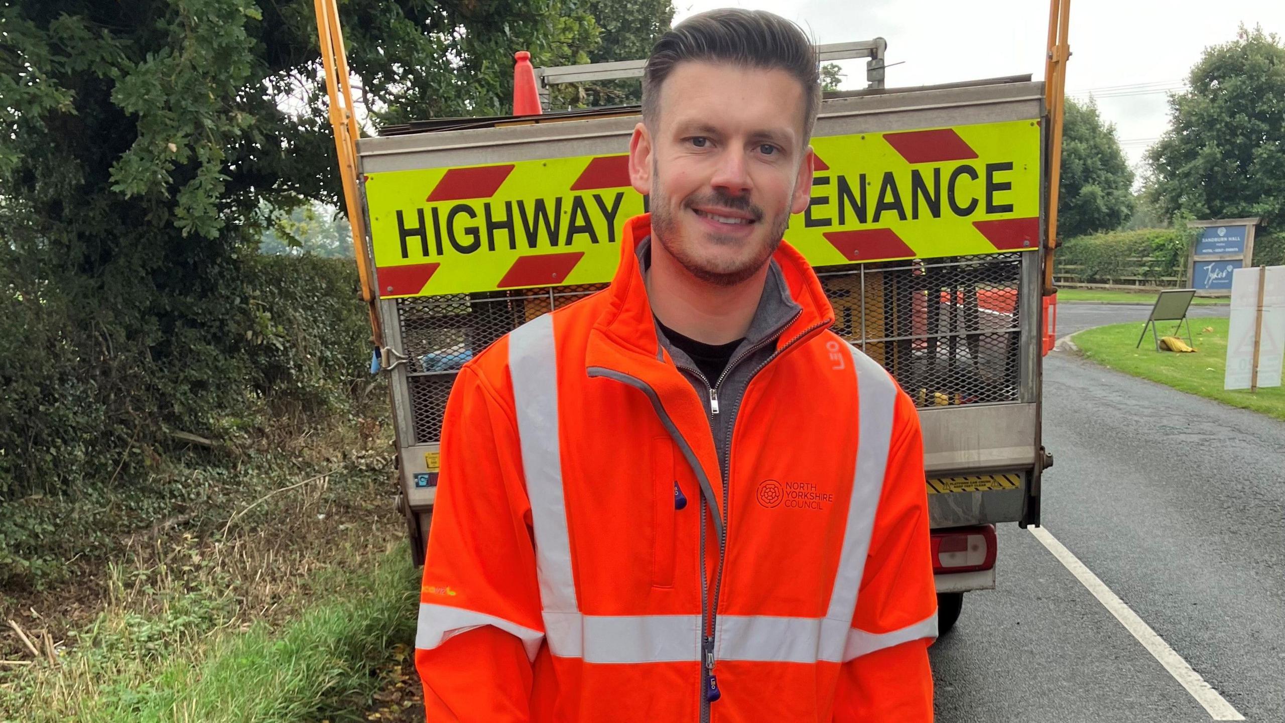 Keane Duncan, from North Yorkshire Council, wears an orange hi-vis jacket and stands in front of a highway maintenance vehicle