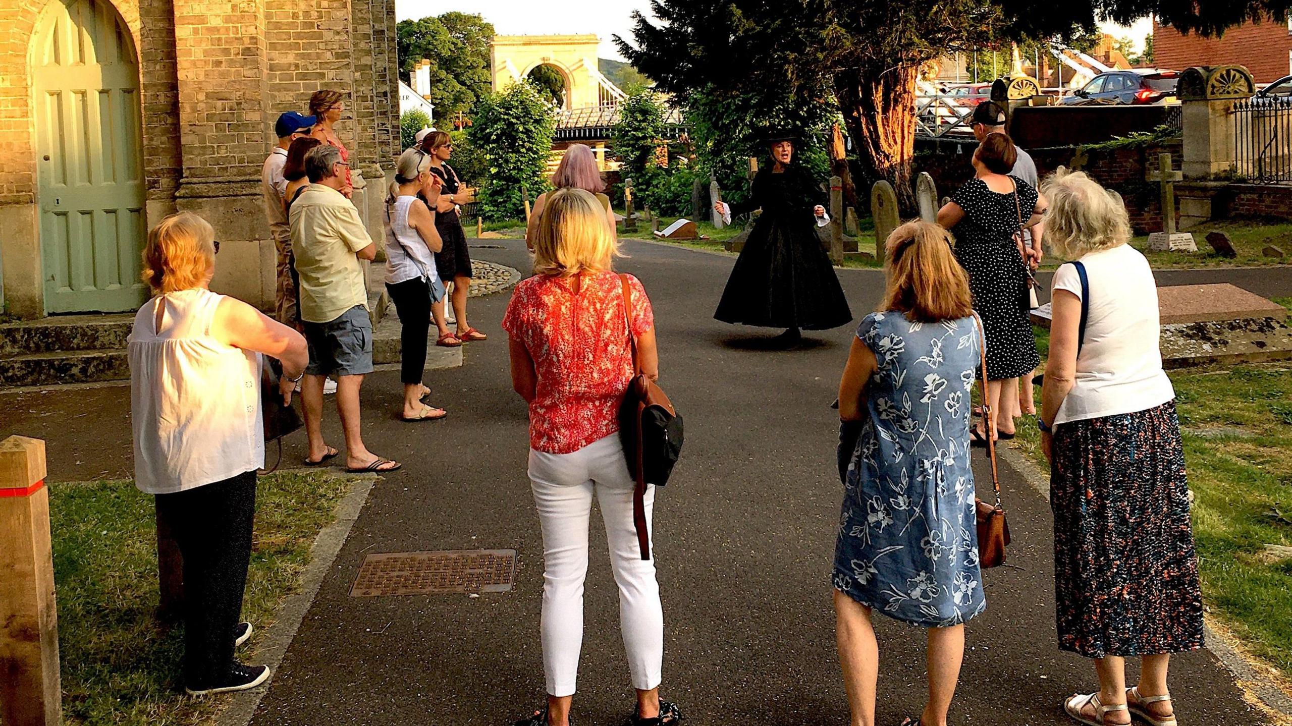 A woman dressed in all black clothing from the 1800s, is speaking to a crowd of people wearing modern summer clothes.