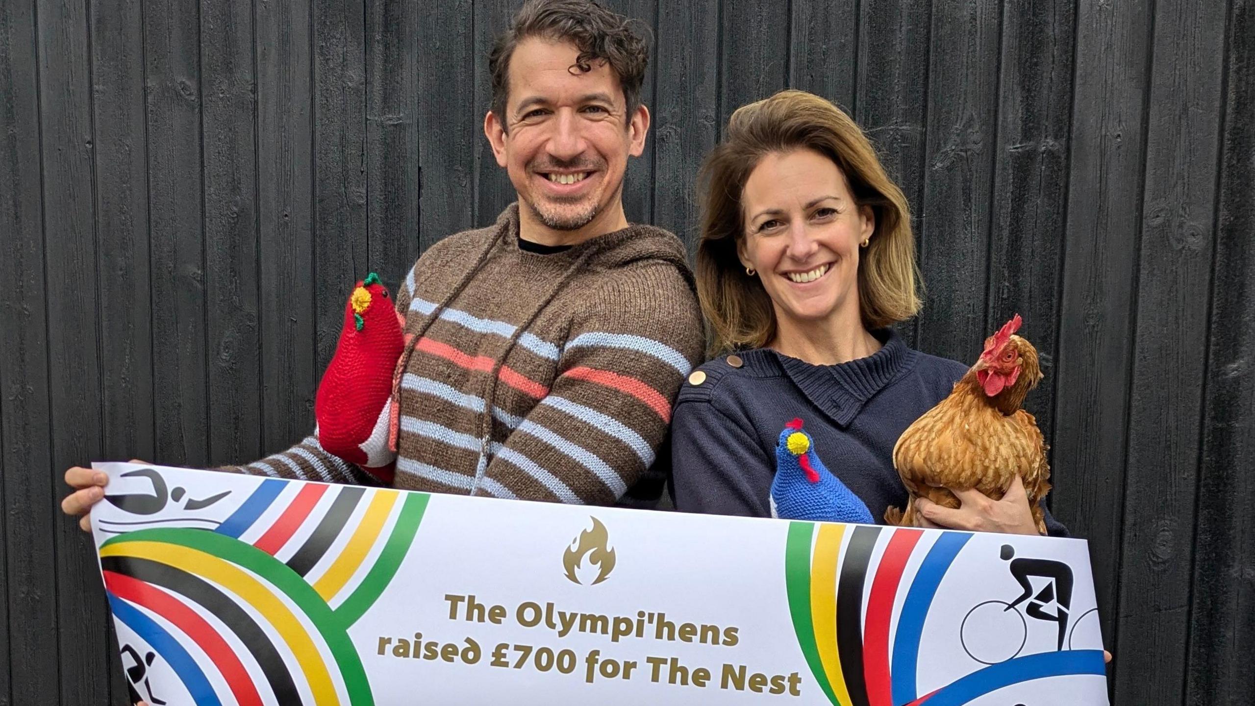 Stuart Race is pictured standing next to The Nest founder Hannah Tuckwell. They are holding a banner that reads: The Olympi'hens raised £700 for The Nest. They are both smiling at the camera while Ms Tuckwell is holding a real chicken and Mr Race holds one of the knitted hens.